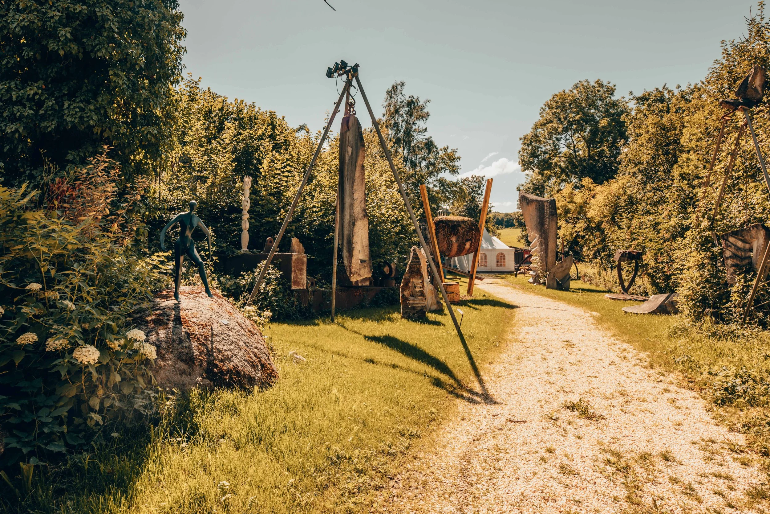 Skulpturenausstellung im Freien auf einem grünen Rasenweg, umgeben von Bäumen und einer weissen Zelt im Hintergrund.