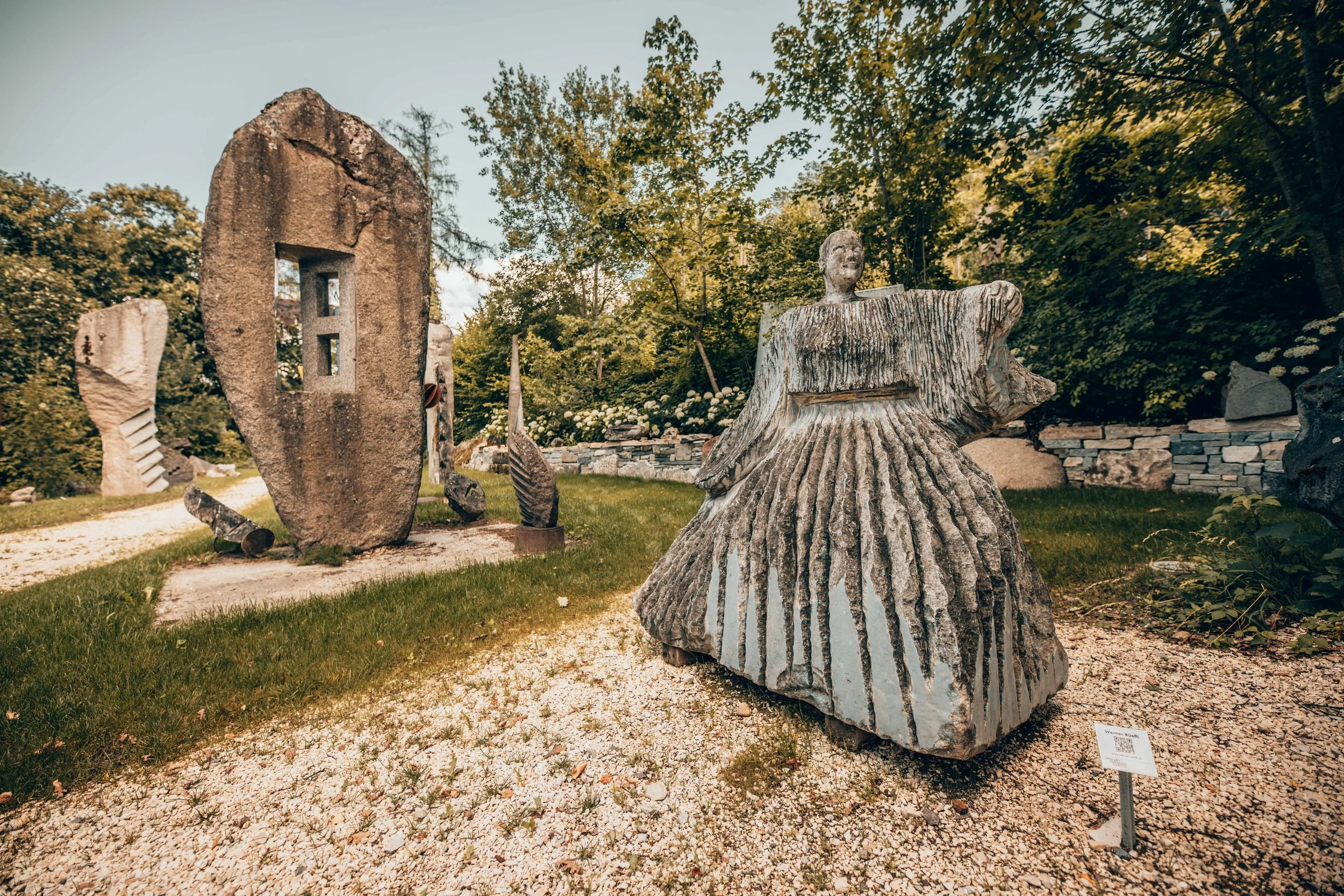 Skulpturen im Garten, darunter die eines Mädchens in einem Kleid, umgeben von weiteren Steinskulpturen und Pflanzen, bei Tageslicht mit blauen Himmel.
