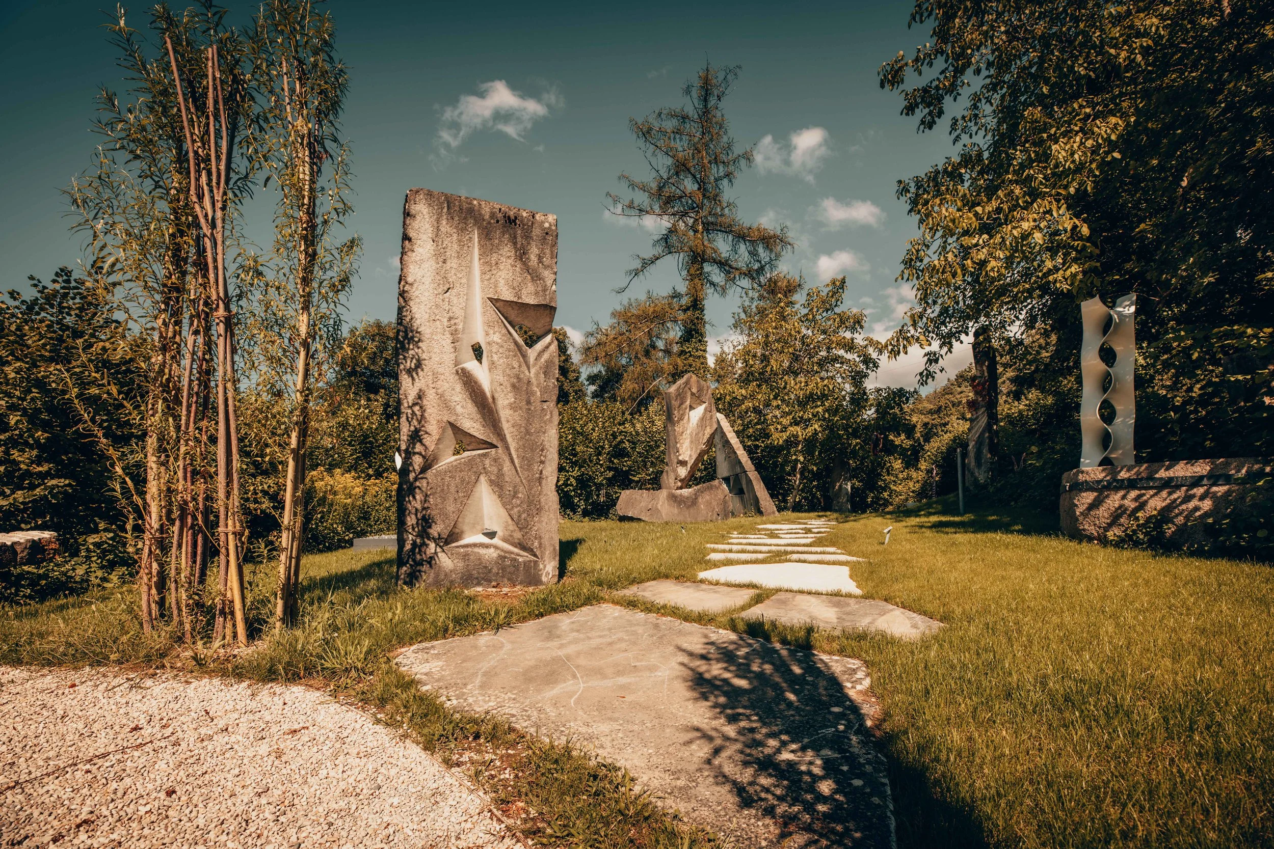 Kunstwerke im Garten, bestehend aus mehreren grossen Skulpturen aus Stein und Metall, umgeben von Bäumen und Rasen, bei sonnigem Himmel.
