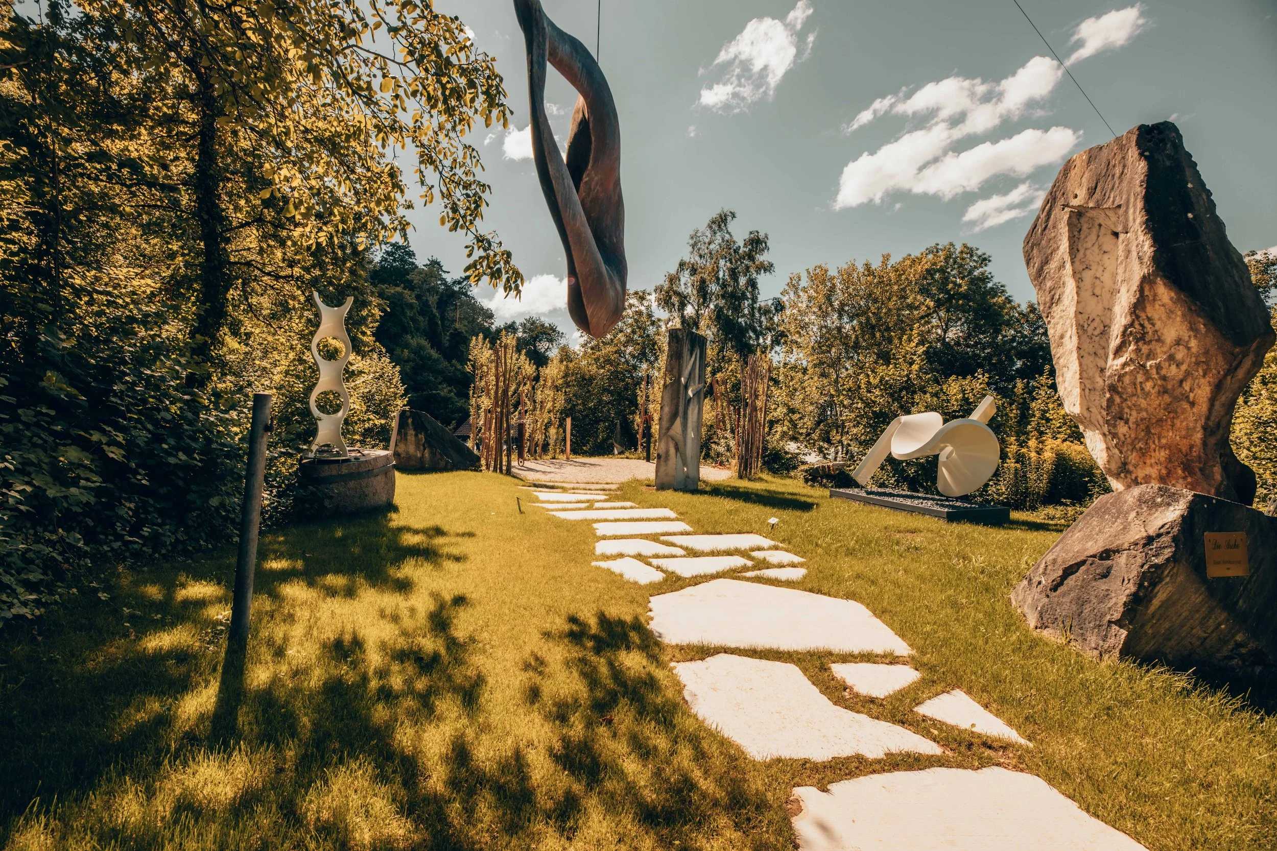 Kunst im Freien mit Steinskulpturen und einem Weg aus grossen, unregelmässigen Steinen, umgeben von Bäumen und Blättern unter einem bewölkten Himmel.