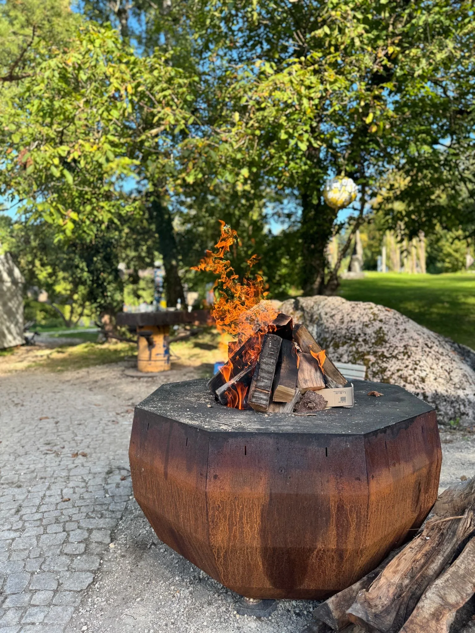 Feuer brennt in einer grossen Metalltonne im Freien, umgeben von Bäumen und Gras, bei schönem Wetter.