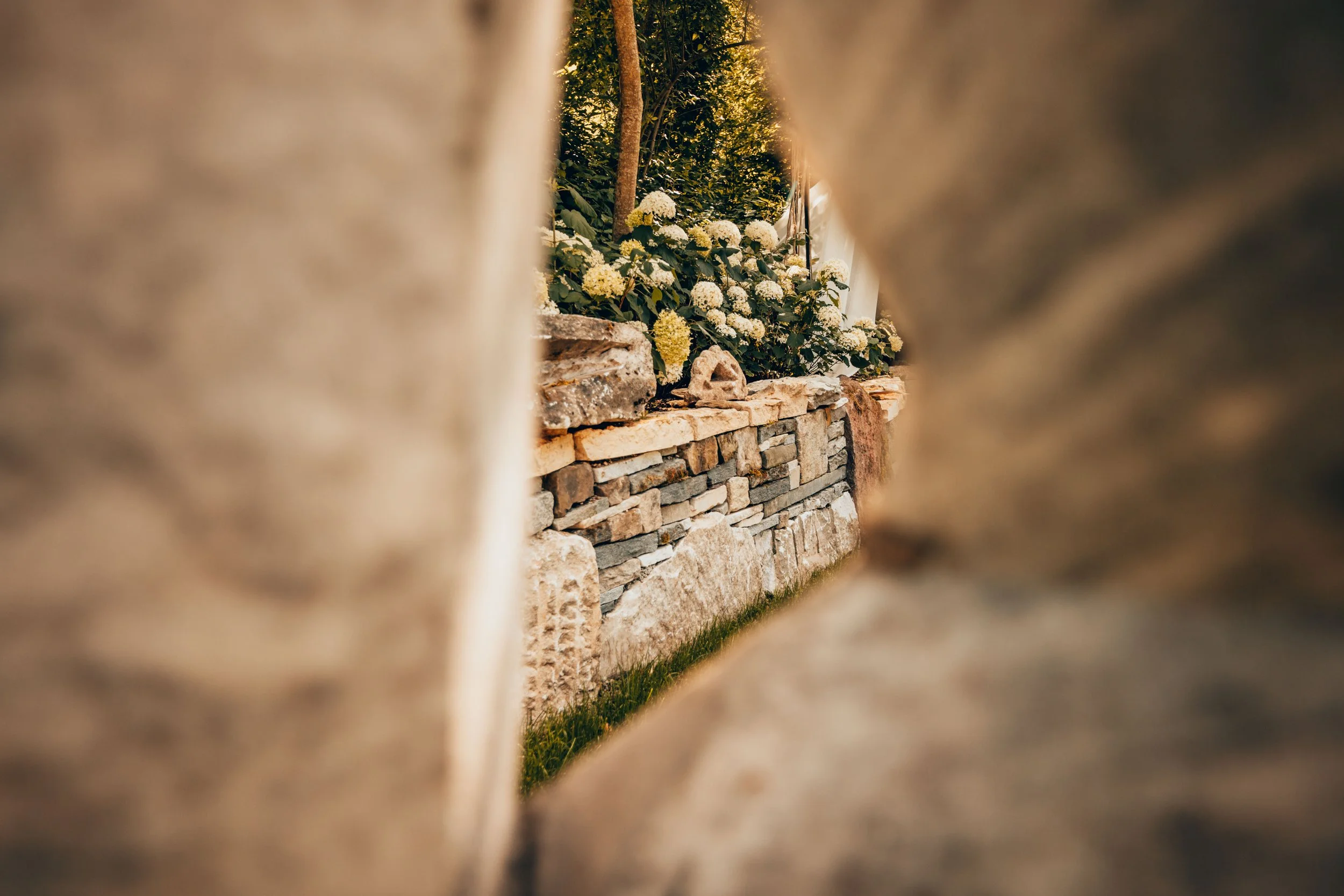 Blick durch eine kleine Lücke zwischen zwei Steinen auf einen Garten mit einer Steinmauer und weissen Blumen.