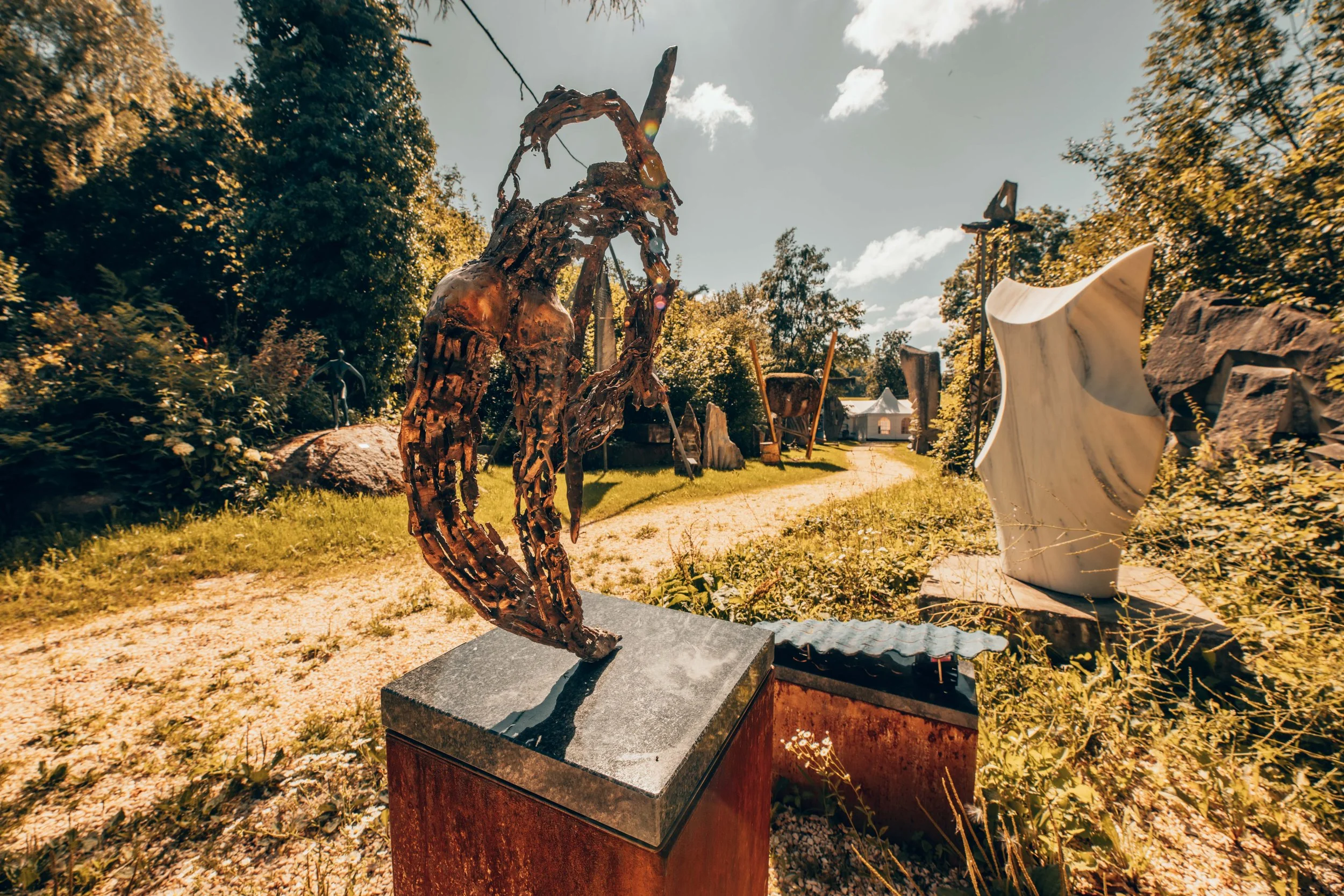 Kunstinstallation im Freien, bestehend aus einer Metallskulptur eines im Detail ausgearbeiteten, stilisierten Vogels und einer weiteren abstrakten Skulptur aus Mist and Stein, auf einem sonnigen Tag in einem Kunstgarten mit Bäumen im Hintergrund.