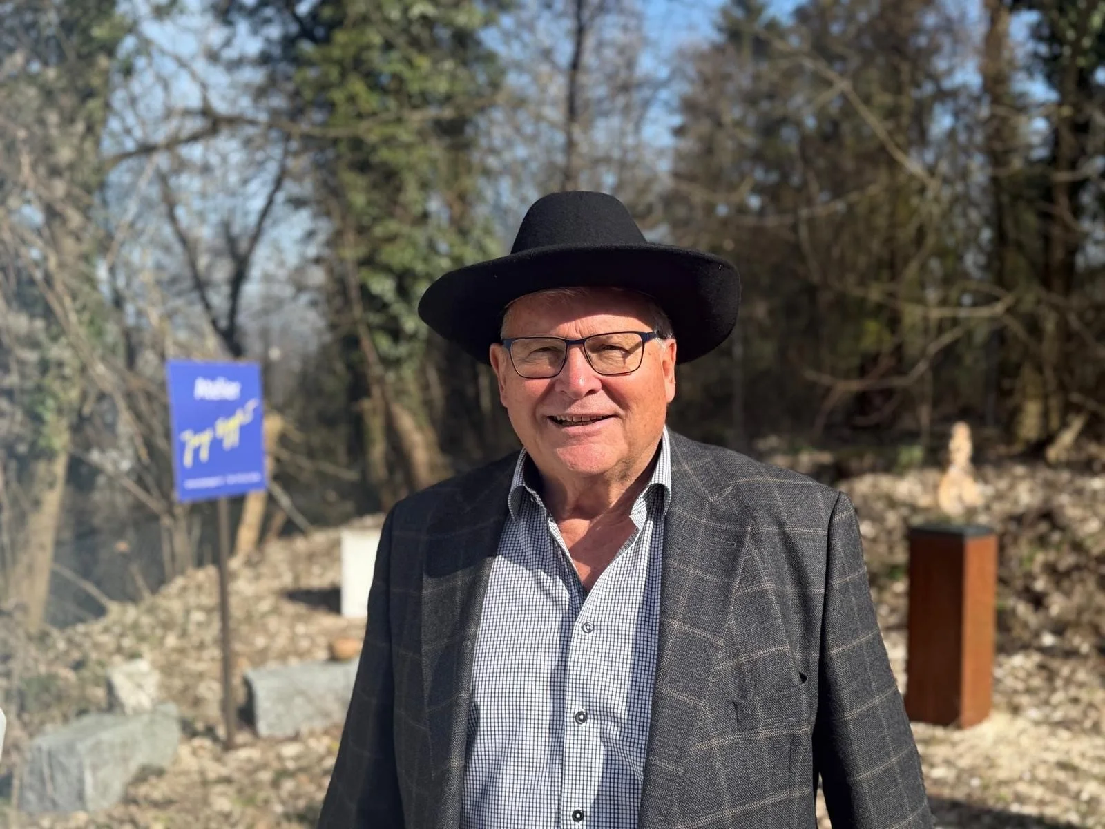 Jorge Egger trägt einen schwarzen Hut, eine graue kariert Jacke und ein Hemd. Er steht im Freien mit Bäumen im Hintergrund.