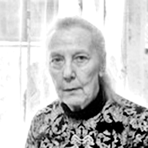 Schwarz-weiß-Foto einer älteren Frau mit kurzem, hellen Haar, trägt ein floral gemustertes Oberteil und sitzt vor einem vertikalen Holzhintergrund.