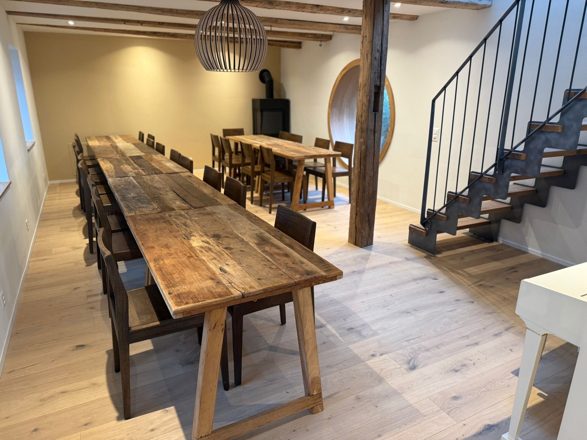 Ein Esszimmer mit langen Holztischen und Stühlen, Holzdecke, Stützpfeiler, offenes Stiegenhaus mit schwarzen Metallgeländern, ein Kamin im Hintergrund.