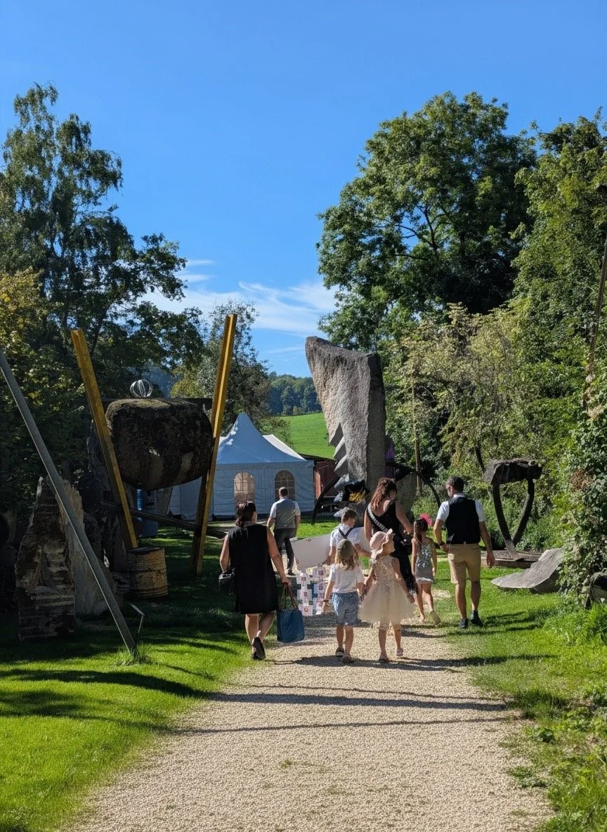 Menschen spazieren in einem Aussenmuseum mit grossen Skulpturen aus Holz und Stein, umgeben von Bäumen und einer Wiese.