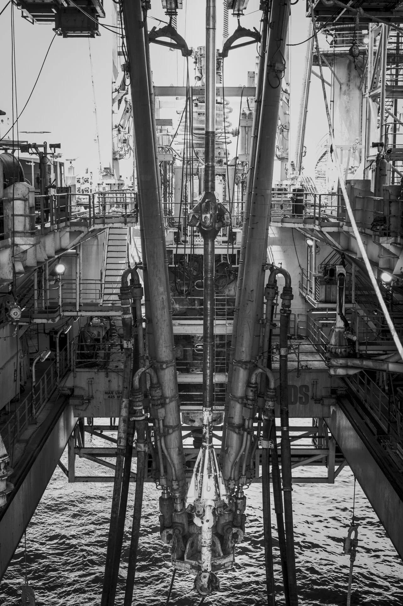 Black and white photo of an offshore oil rig with complex machinery and pipes, viewed from the deck. Water is visible beneath the rig.