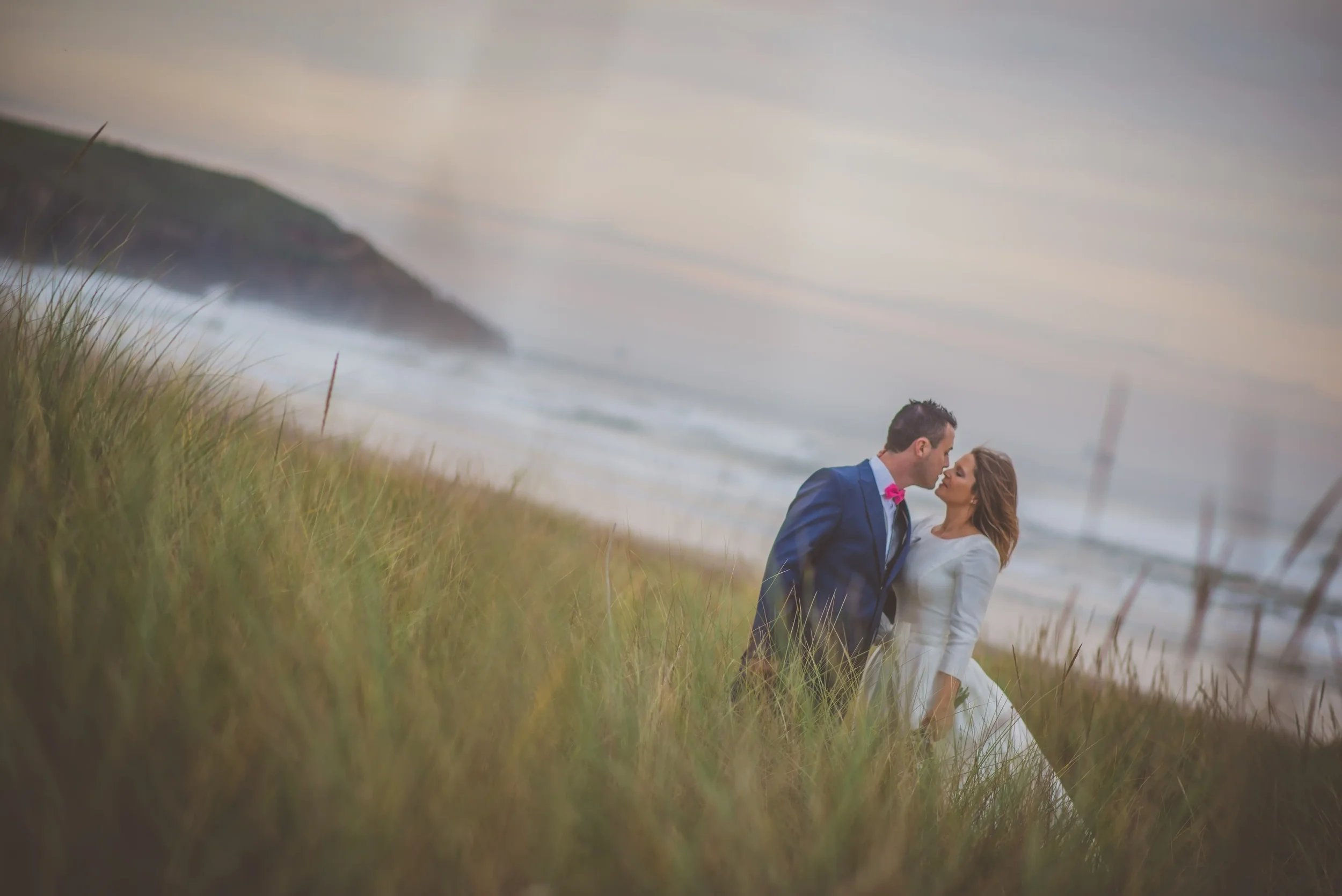 post boda en la playa asturias , posando románticamente al atardecer, con césped y dunas de arena en primer plano y el mar y el cielo en el fondo.