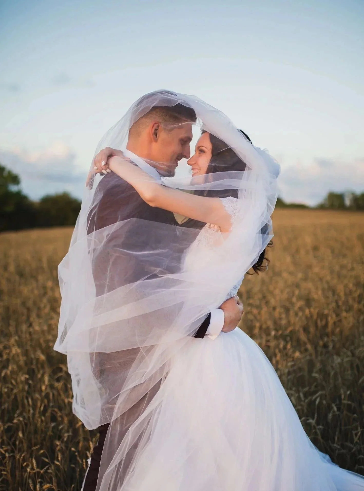 A newlywed couple embracing in a field, with the bride’s veil wrapped around them during sunset.