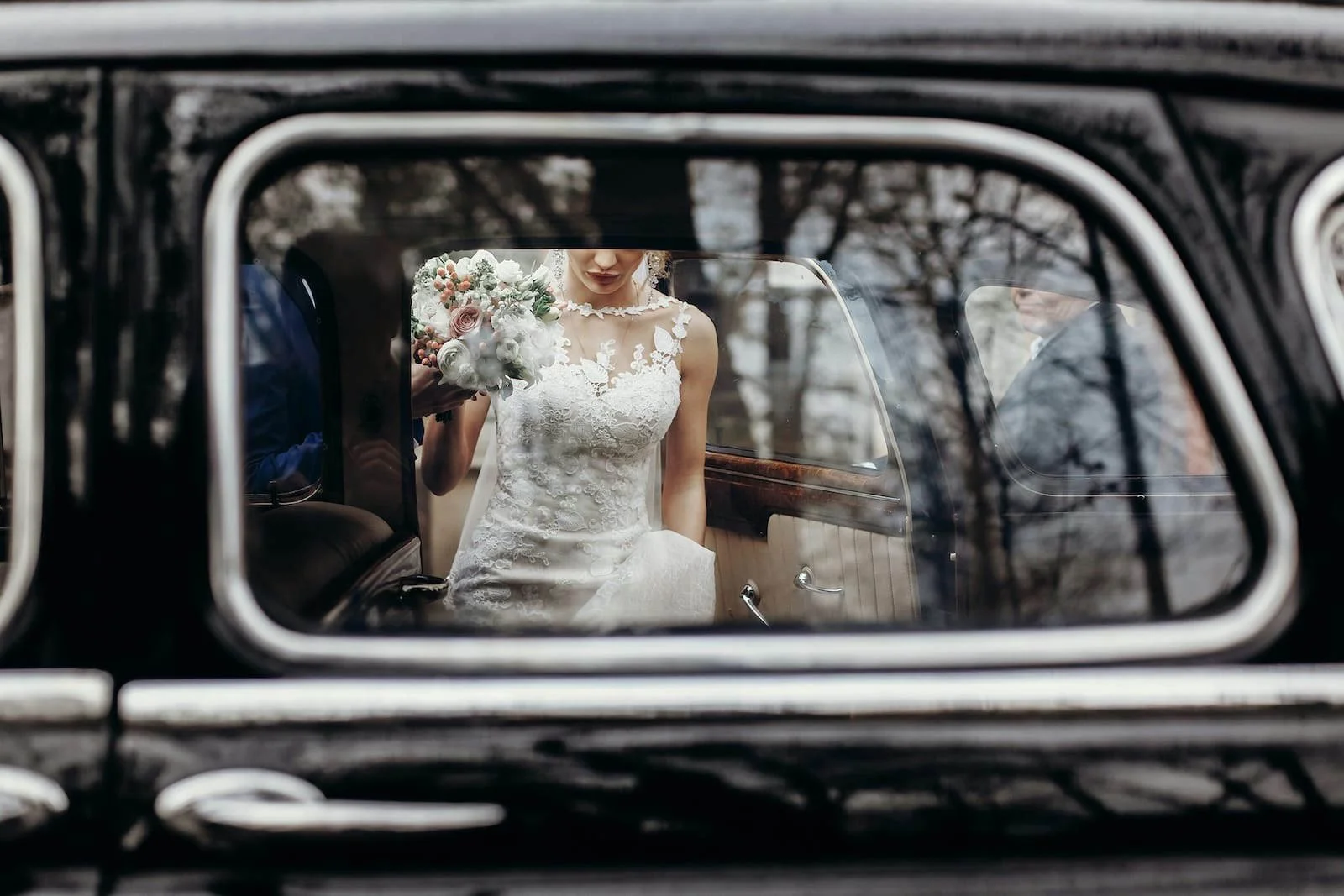 A bride in a white wedding dress holding a bouquet, seen through the window of a vintage black car in a romantic Derbyshire wedding moment