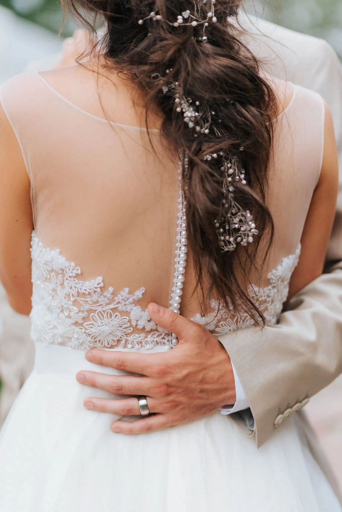 Close-up of a groom’s hand on the bride’s waist, showing the back of her lace wedding dress and intricate hair accessories