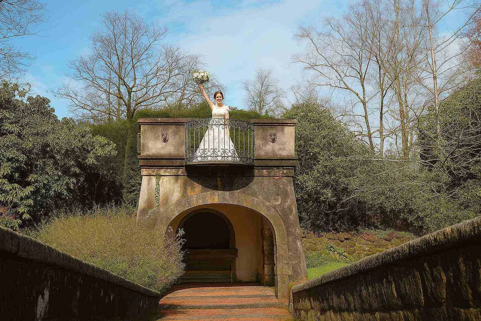Bride holds bouquet aloft on stone balcony at Foxtail Barns gardens