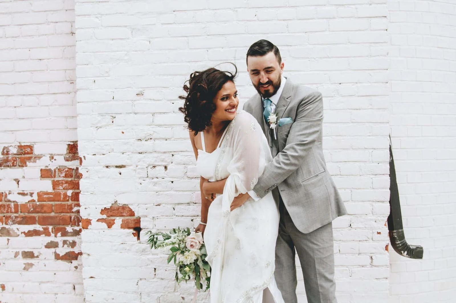 A joyful wedding couple sharing a laugh in front of a whitewashed brick wall