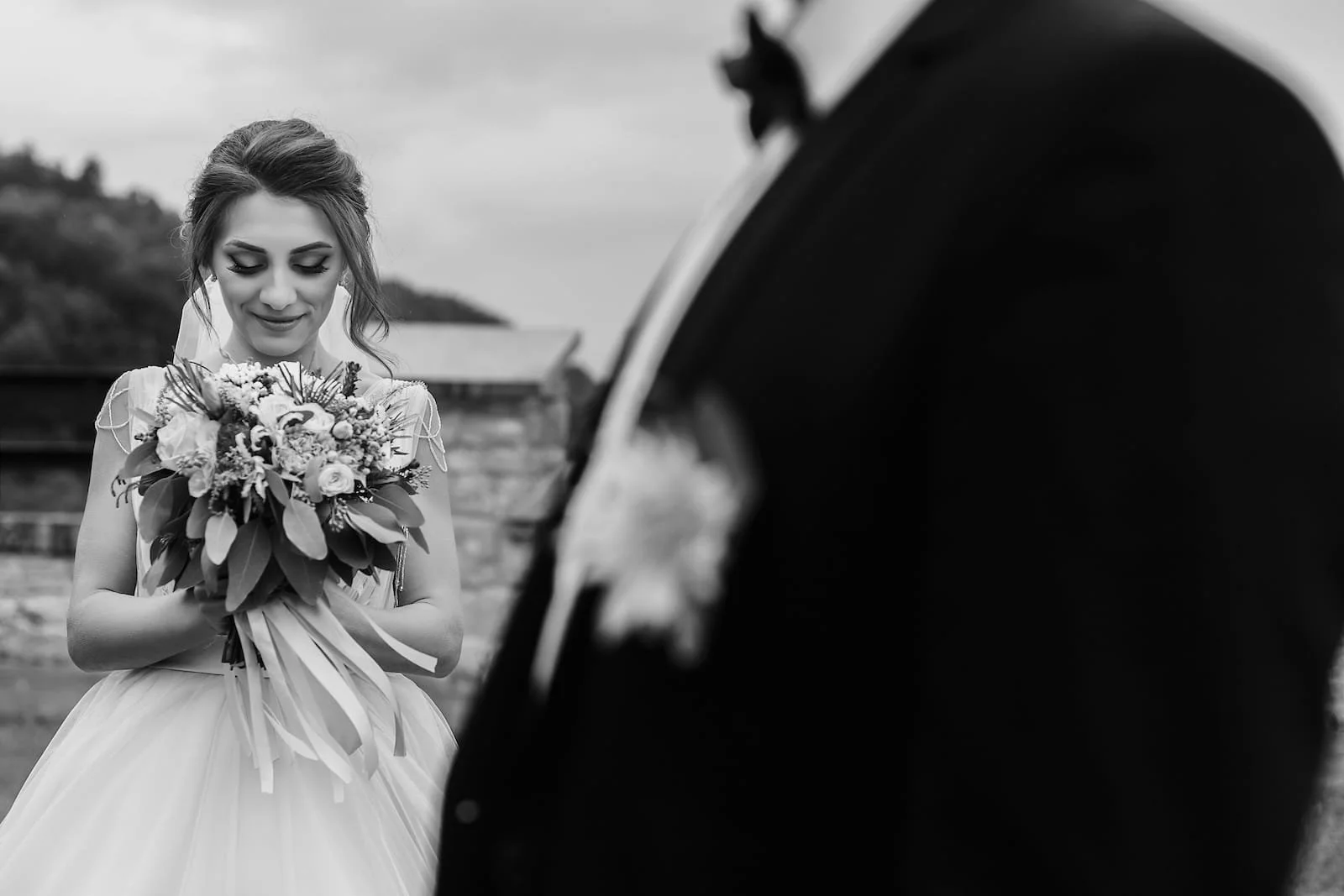 Black and white photo of a bride holding a bouquet, looking down with a gentle smile, and a groom in uniform blurred in the foreground. Outdoor setting with buildings in the background.