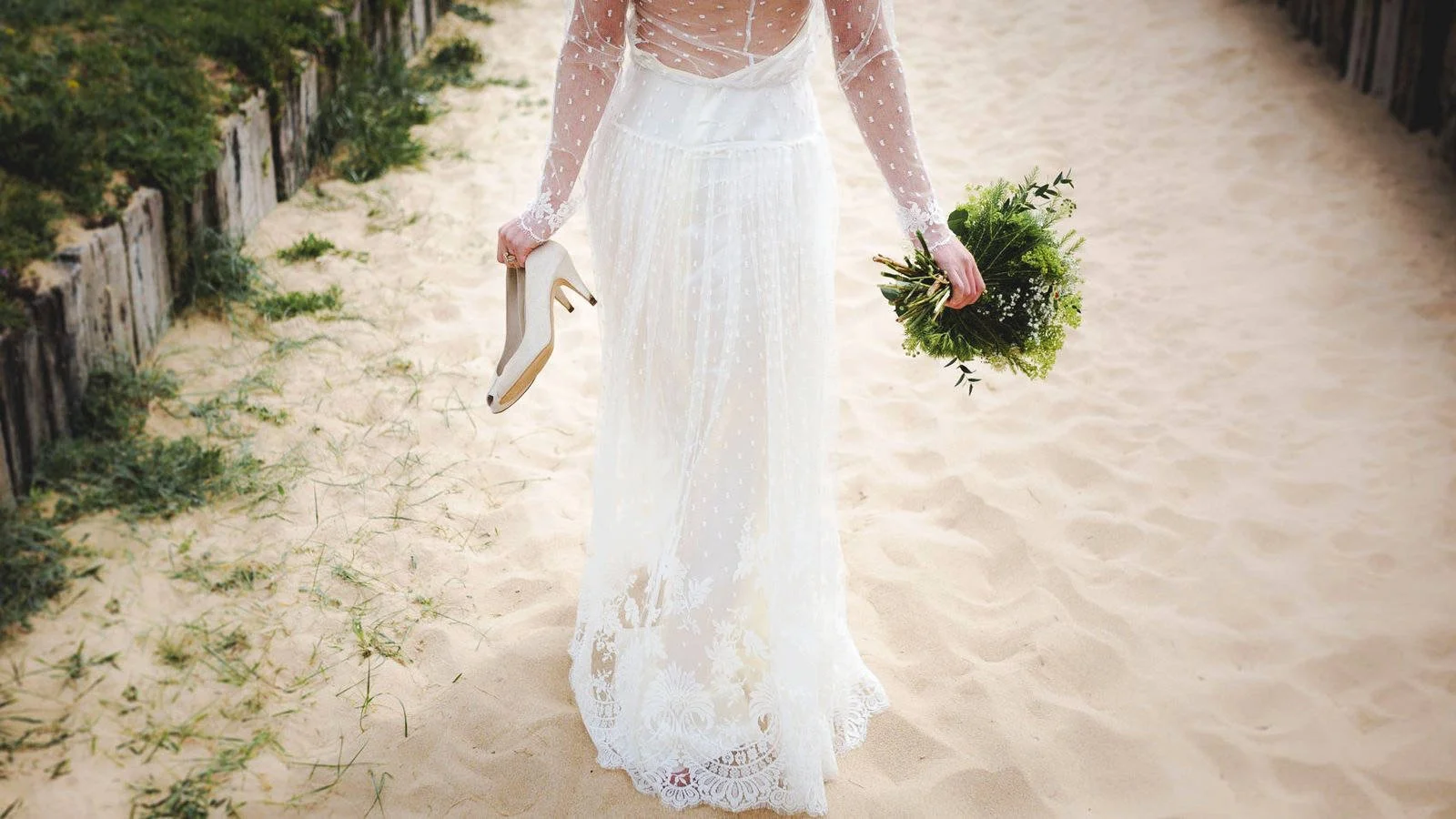 Bride in a white lace wedding dress holding champagne shoes in one hand and a bouquet in the other walking on the beach.