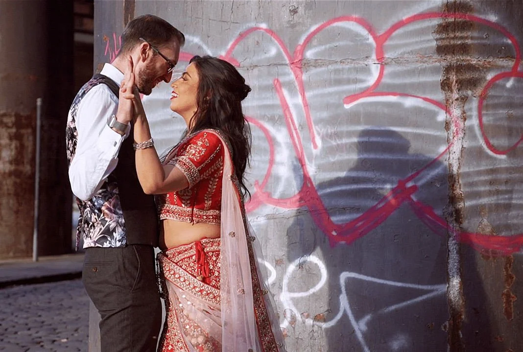 Bride in red lehenga and groom in waistcoat share a joyful moment dancing against a graffiti-covered wall in a vibrant Manchester city centre wedding castlefield