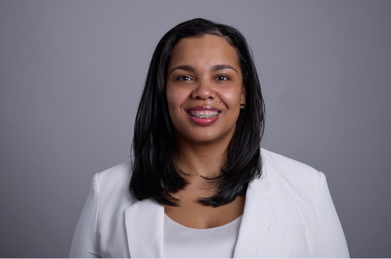 Professional woman with black hair and braces, wearing a white blazer against a gray background.