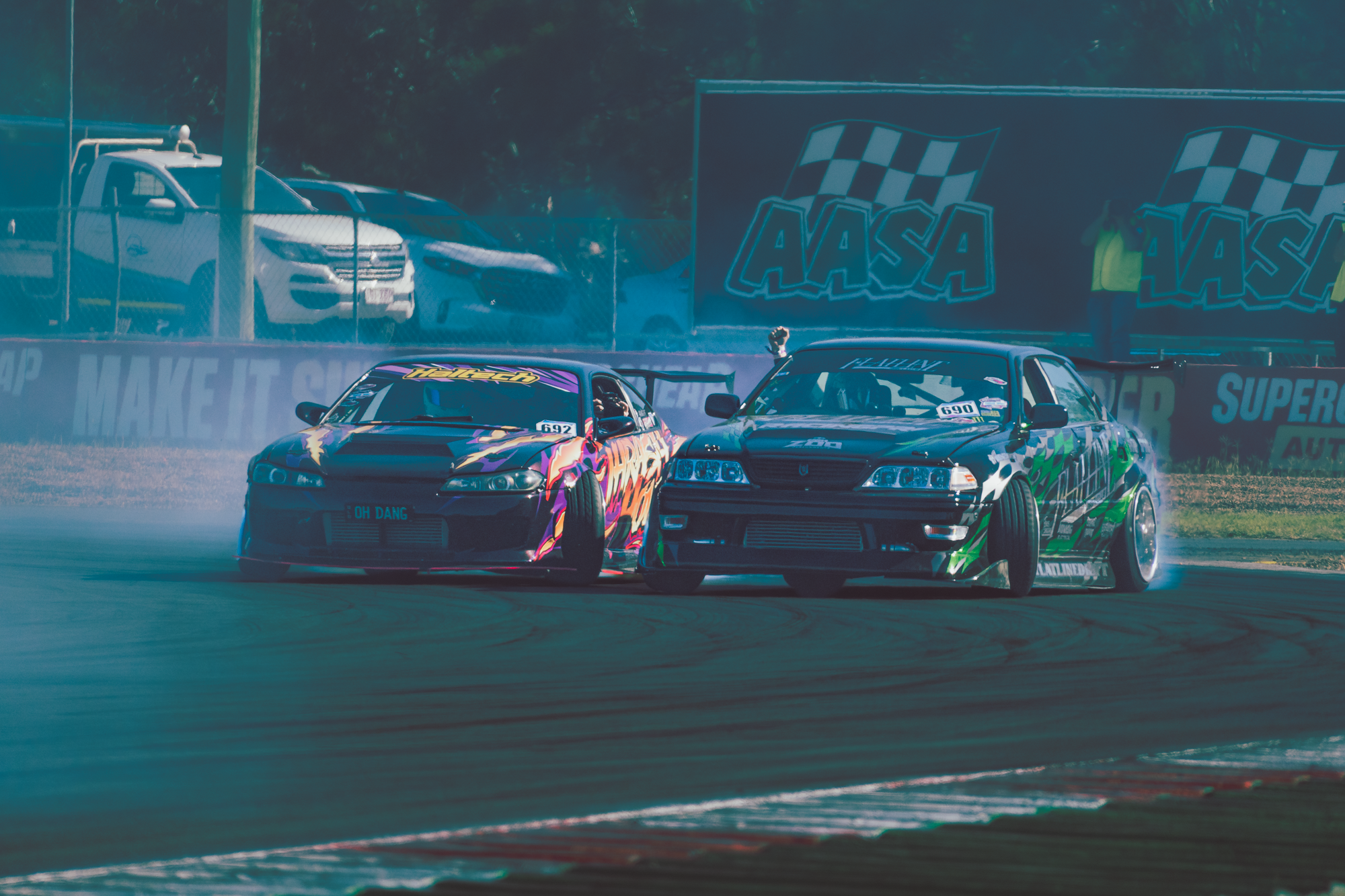 Two race cars competing on a track, one with a colorful lightning bolt design and the other with a green and black design, surrounded by smoke and spectators.