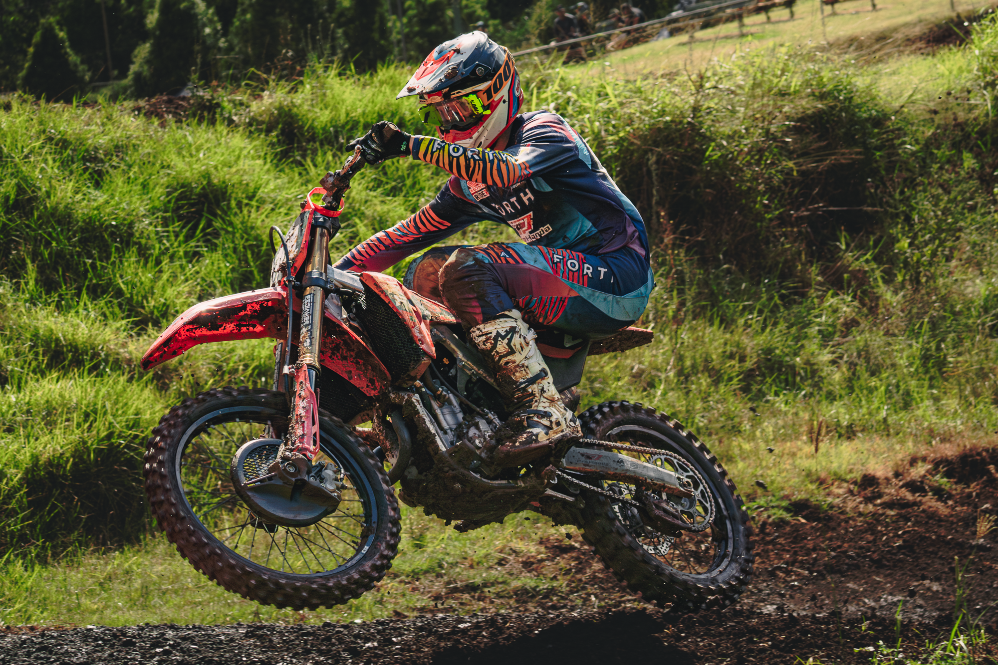 A motocross rider wearing a helmet and colorful gear riding a dirt bike on a grassy track, kicking up dirt.