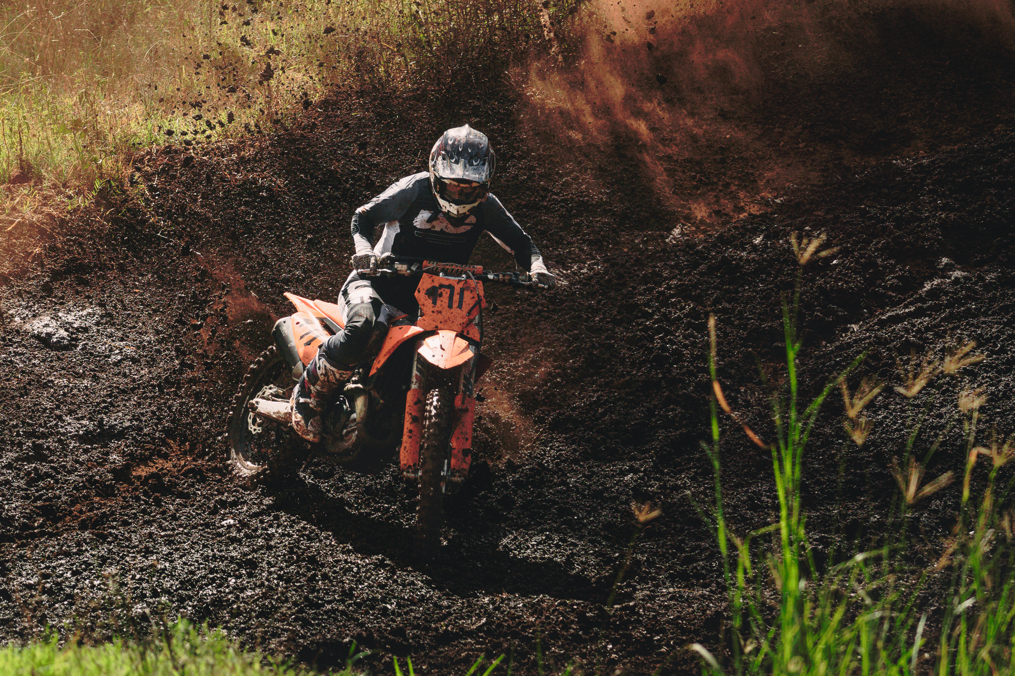 A person riding an orange dirt bike through muddy terrain with mud splashing around, surrounded by green grass and plants.