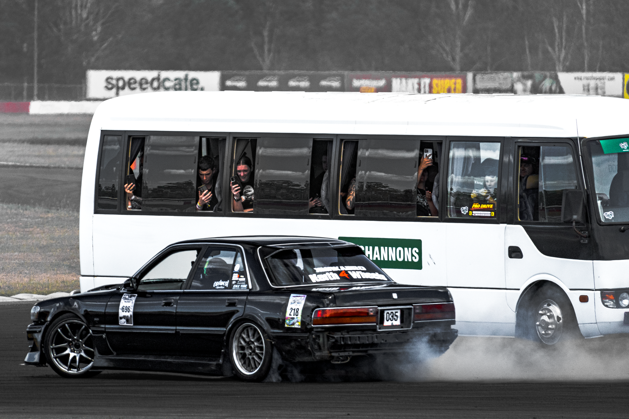 A black sports car drifting on a race track, creating smoke from its tires, with a white passenger bus and overhead billboards in the background.