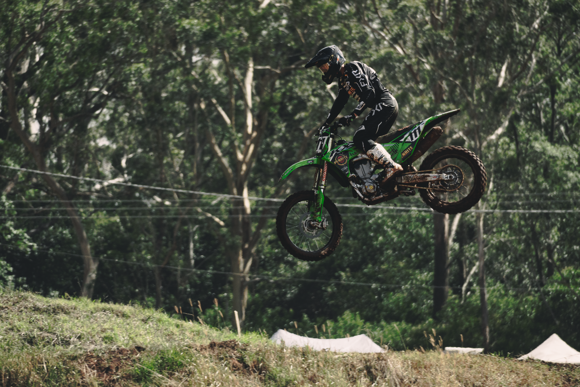 A person wearing a helmet and racing gear riding a green dirt bike off a ramp, with a background of trees and a log jump on a dirt track.