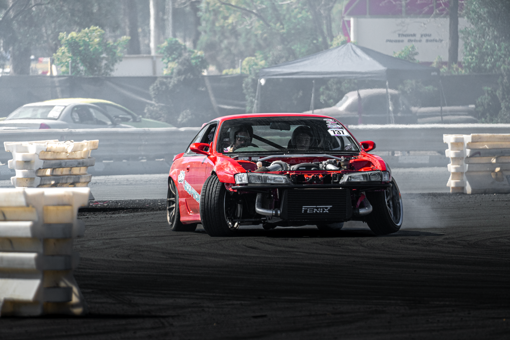 A red race car with the front bumper missing drives on a drag strip, with two people inside. The car is in motion, creating smoke behind it. The background shows a fence, trees, and a tent.