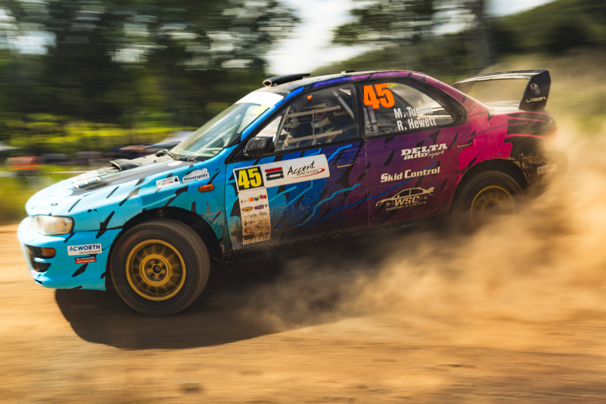 A rally car with the number 45 in orange on its side, speeding on a dirt track with dust behind it. The car is decorated in blue, purple, and black with various sponsor logos, including Delta Auto Sport and Subaru Experts. The background is blurred, 