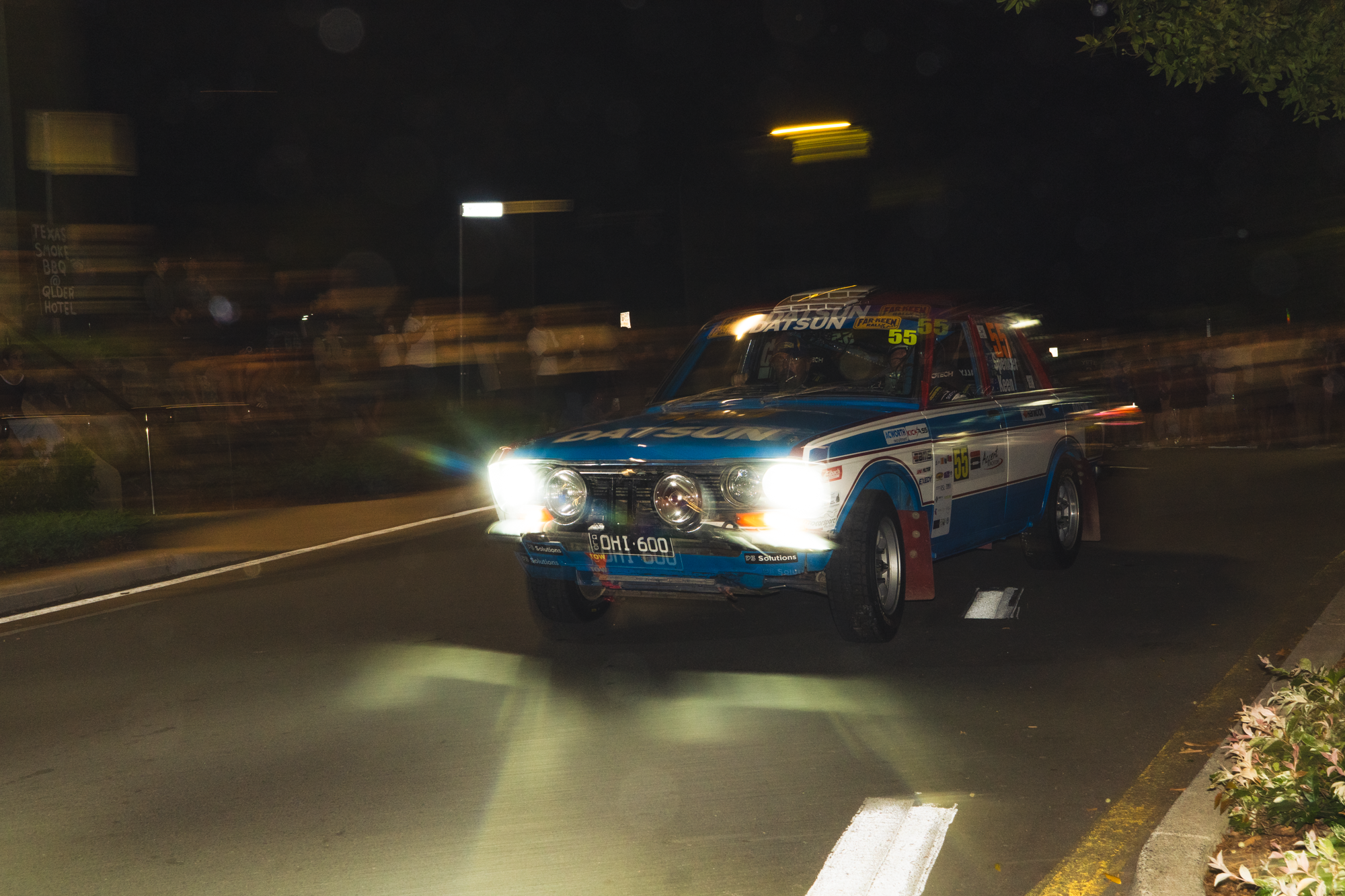 A blue race car with the number 55 on it driving at night, headlights on, on a city street with motion blur and spectators in the background.