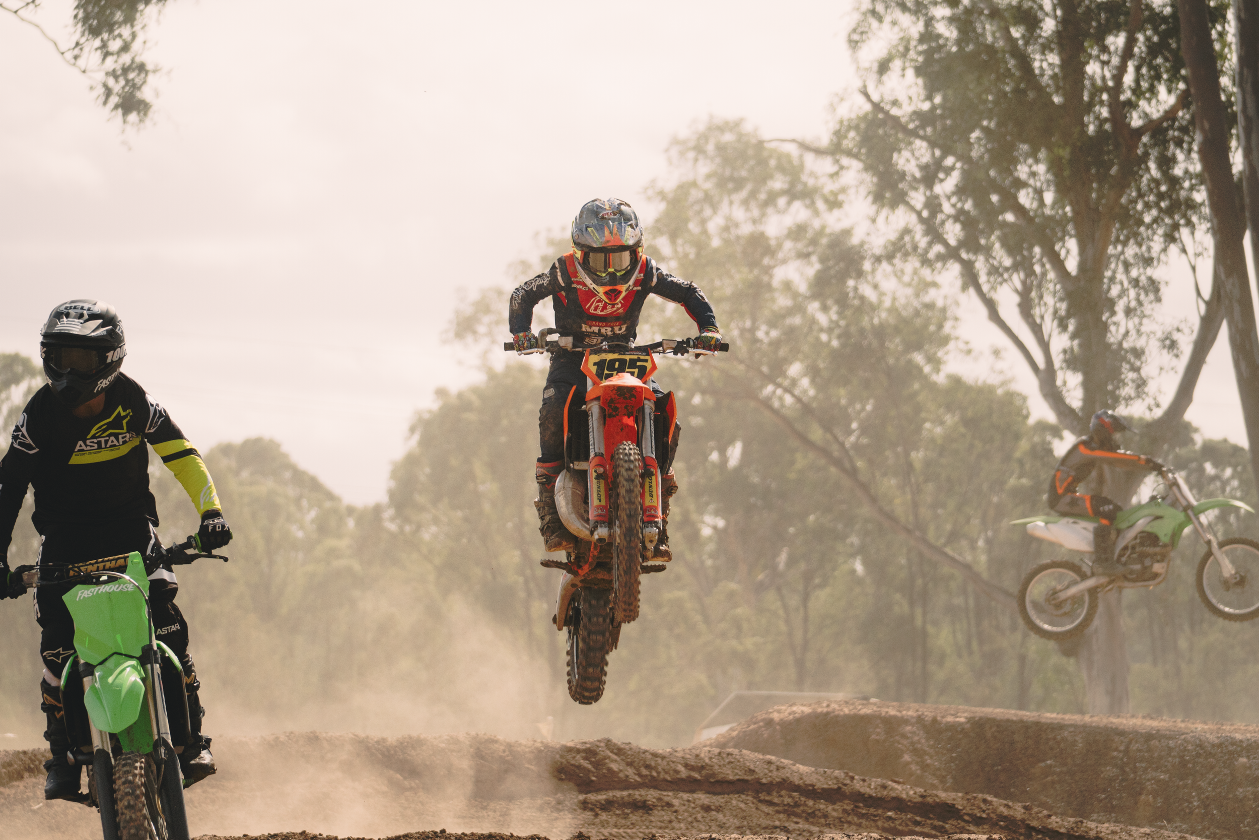 Motocross racers on dirt track with one rider airborne and others on the ground, trees in background.