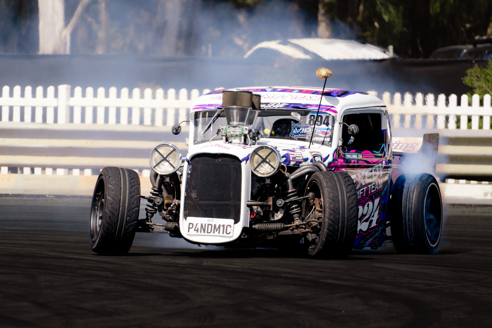 A racing car on a track, kicking up smoke, with white fencing and trees in the background.