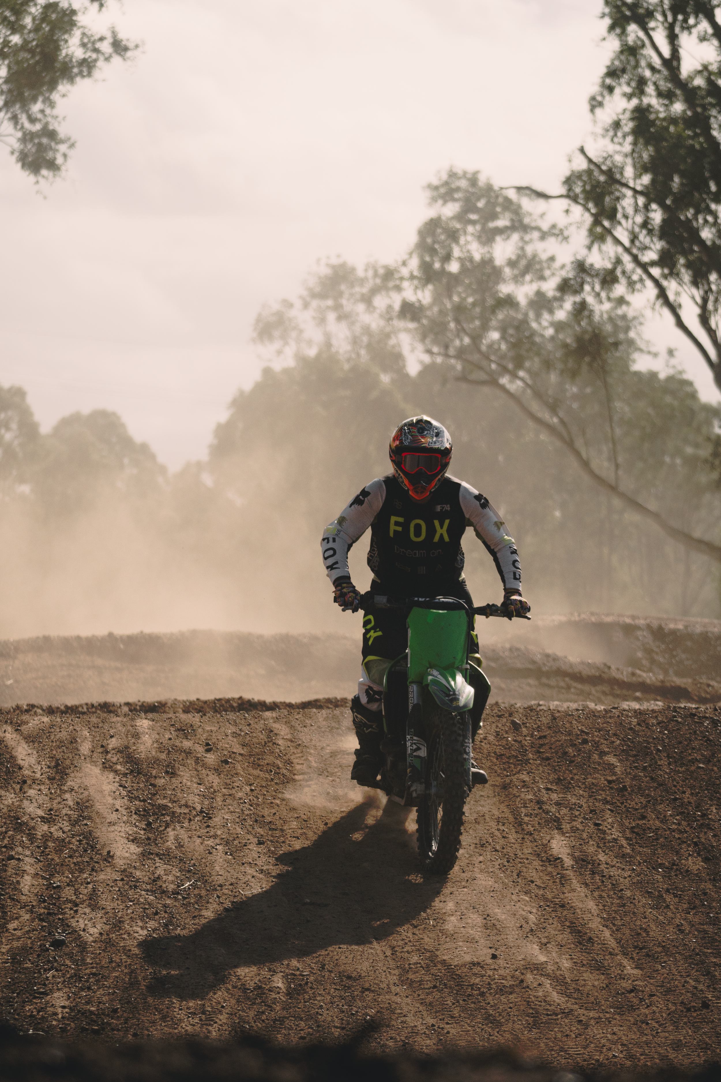A person riding a dirt bike on a dusty trail surrounded by trees, with dust and dirt kicking up behind them.