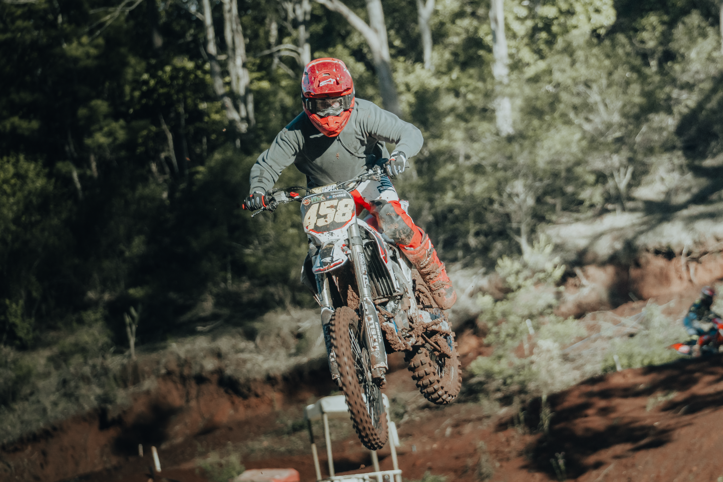 A motocross rider wearing a red helmet and gray protective gear riding a dirt bike on a wooded trail.