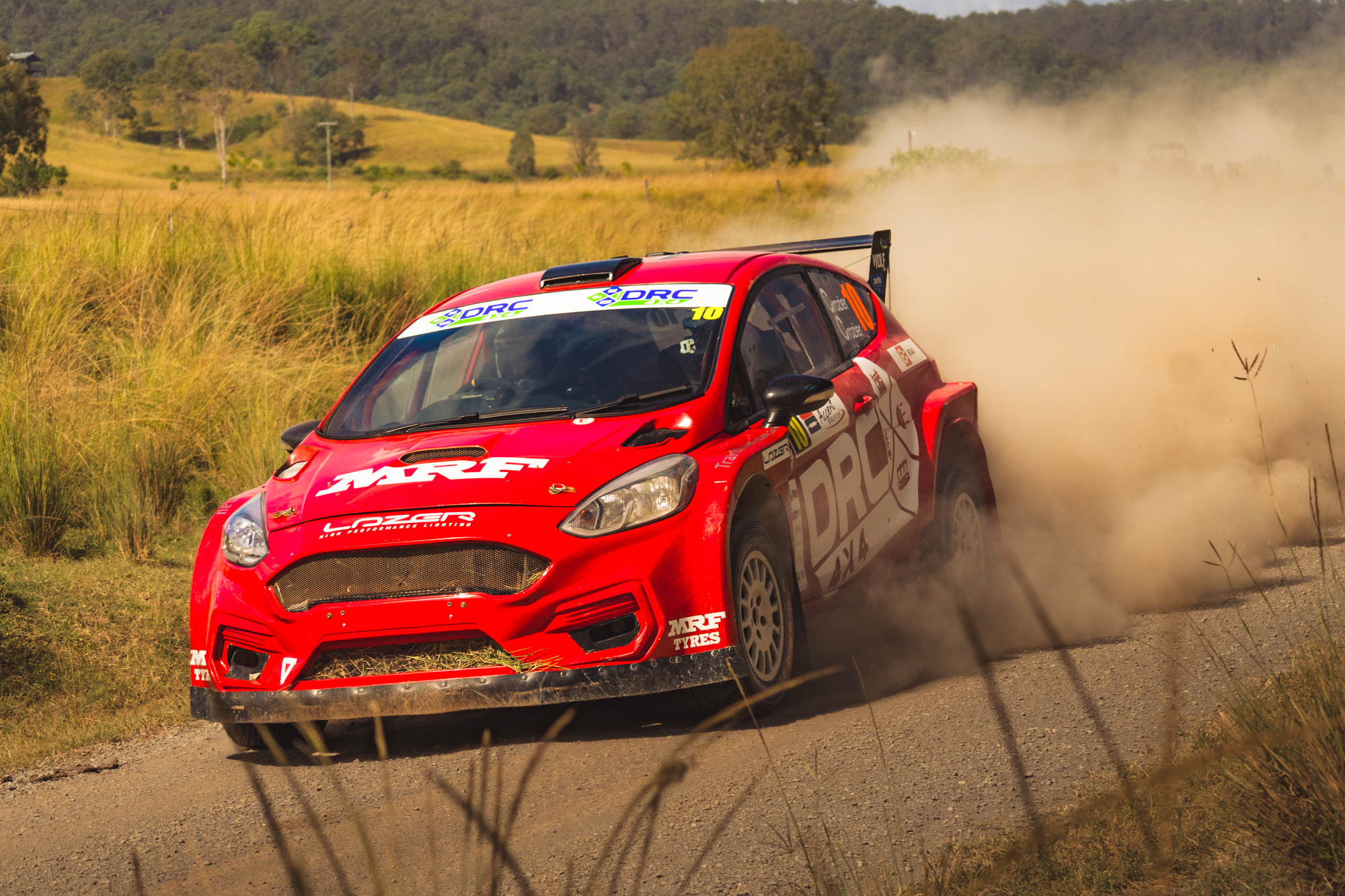 Red rally car speeding on dirt road, kicking up dust, with a rural landscape of grassy fields and trees in the background.