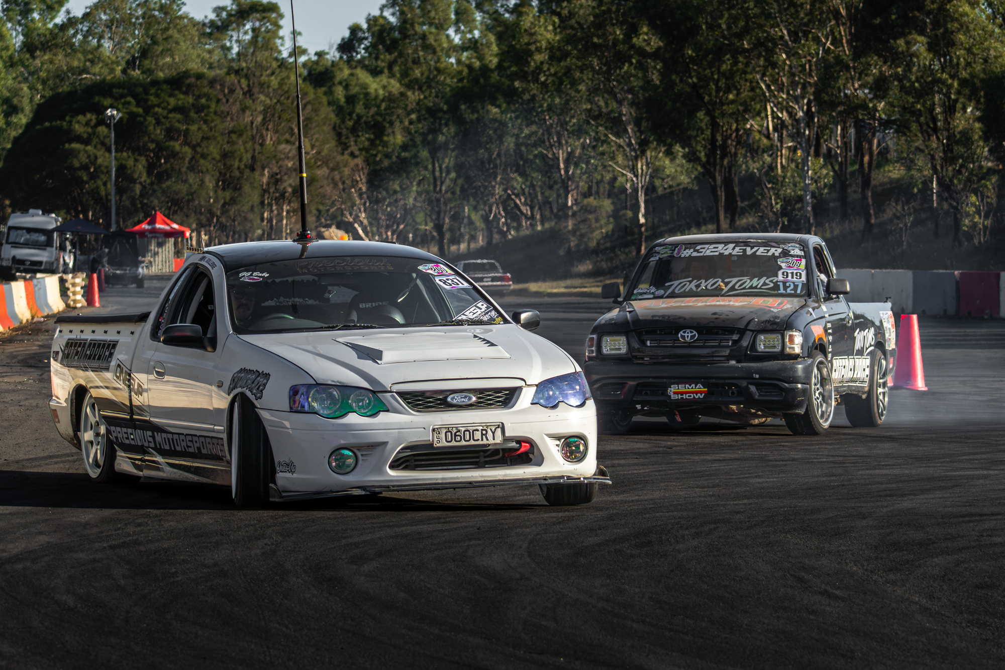 Two race cars driving on a drag strip, with trees and tents in the background.