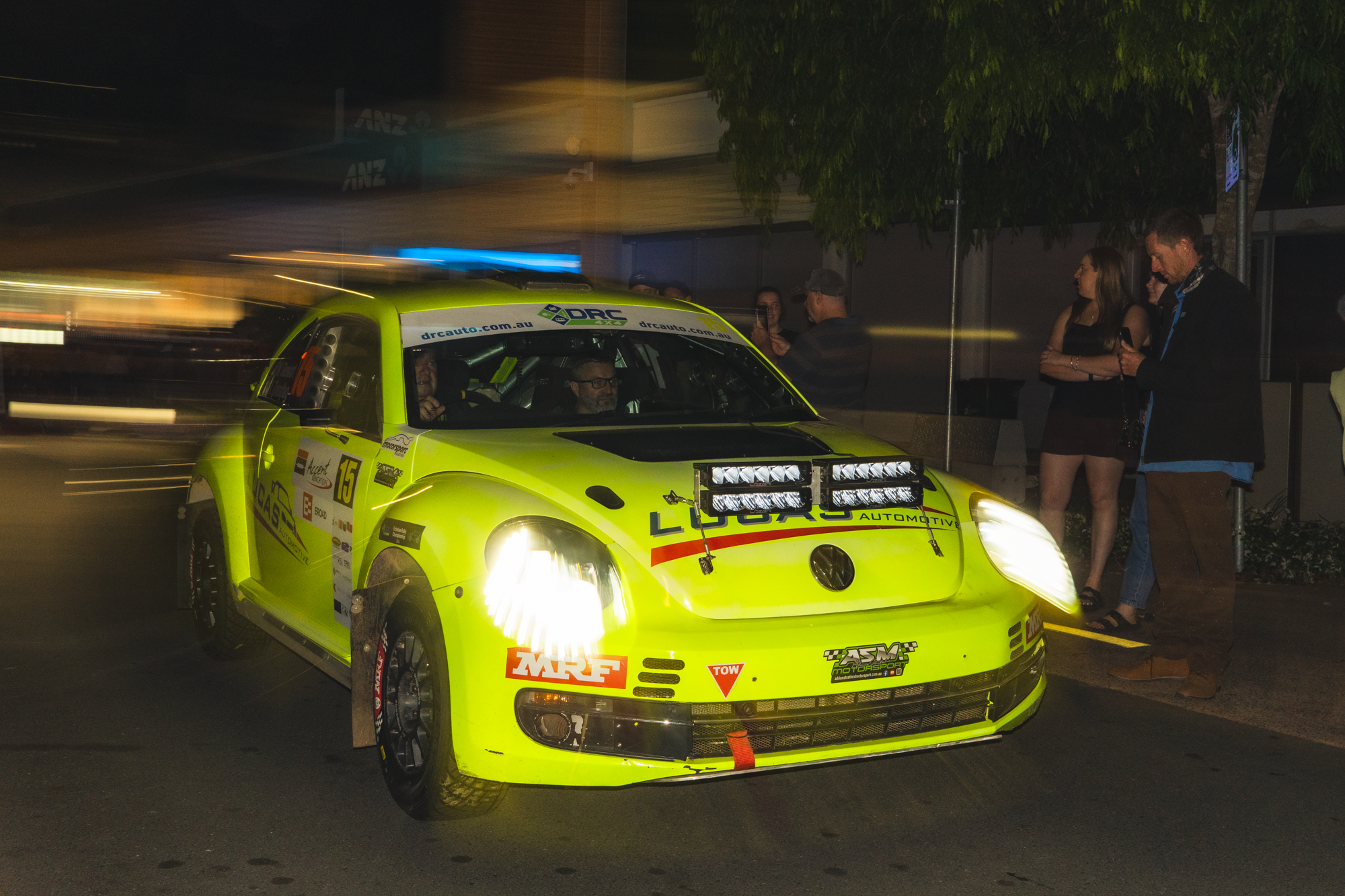 Bright yellow race car with various sponsor logos and bright headlights, parked at night with people standing nearby.