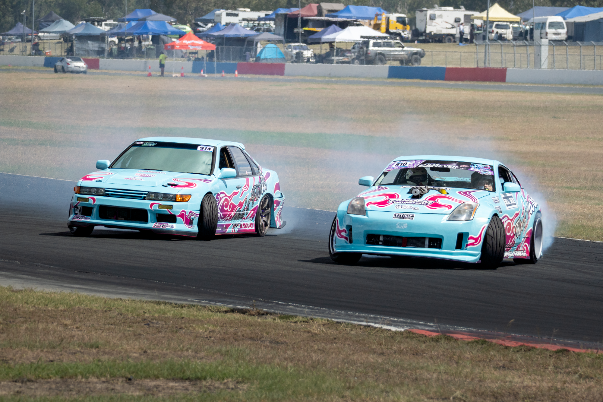 Two blue race cars with pink and white flame decals racing on a track, kicking up smoke, with tents and vehicles in the background.