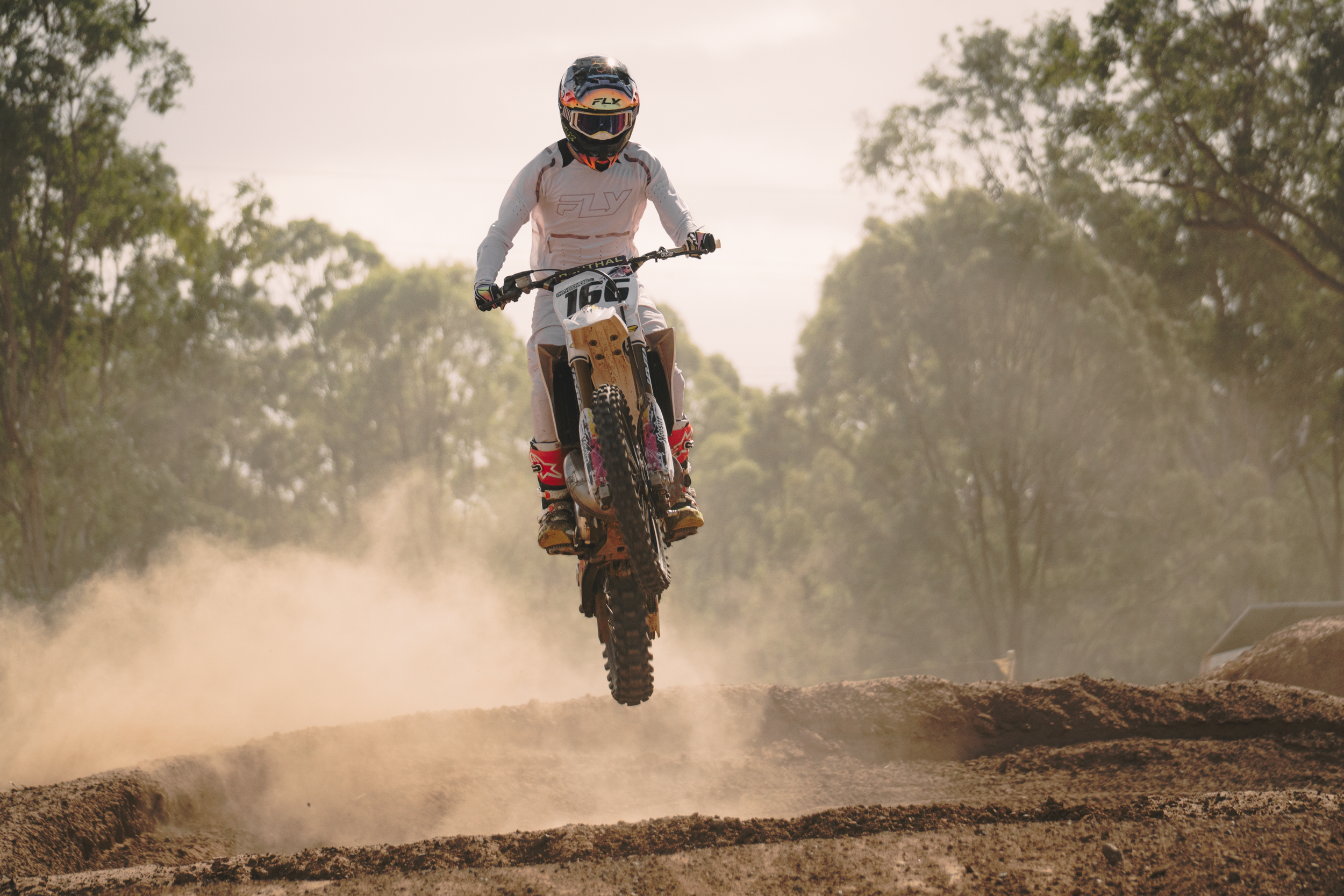 Motocross rider in a white suit and black helmet, riding a dirt bike over a dirt track, kicking up dust, surrounded by green trees.