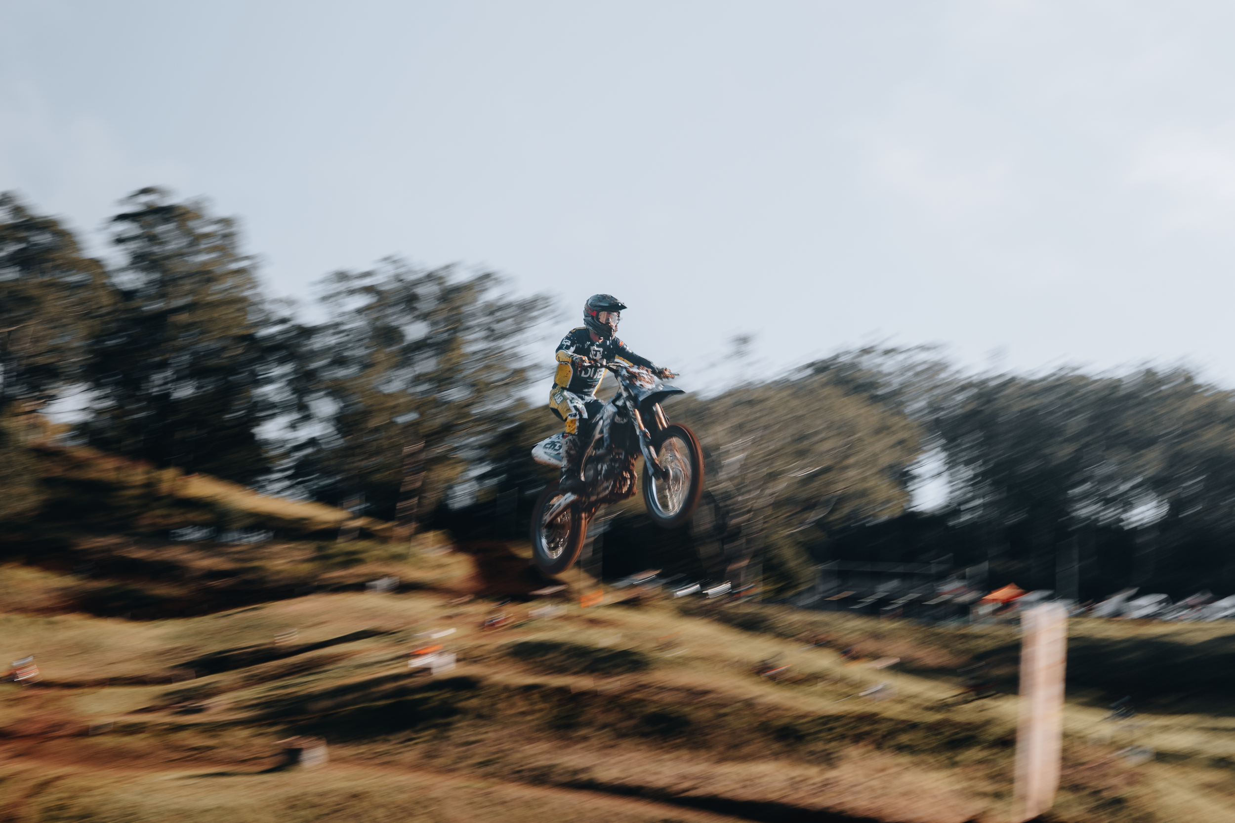 A person wearing a helmet and riding gear is riding a dirt bike in mid-air during a jump on a dirt track, surrounded by trees.