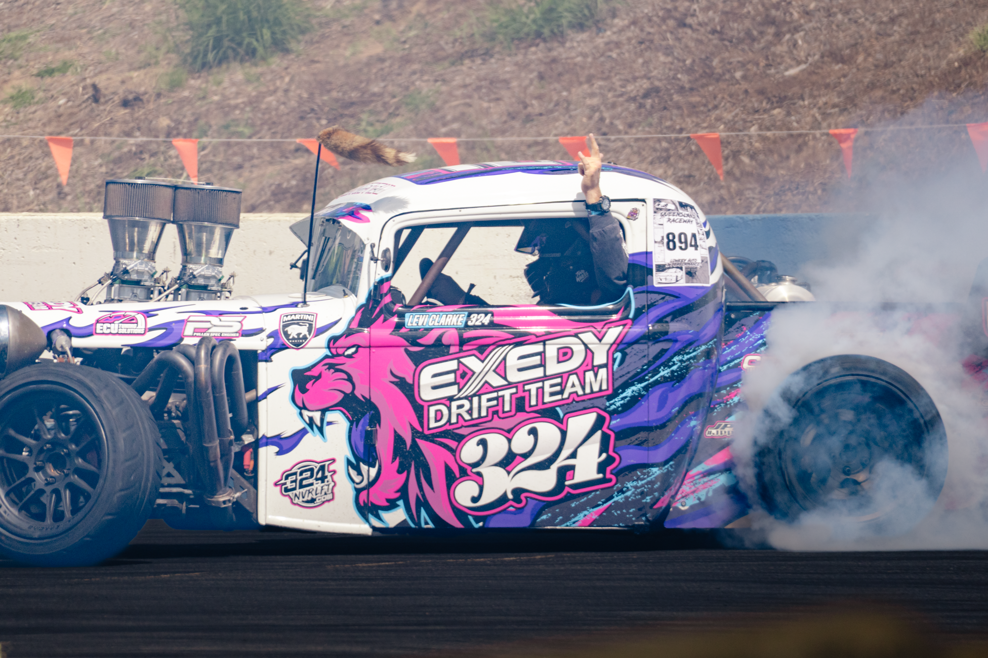 A drag racing car with vibrant pink and purple graphics, showing a man with his hand raised in a peace sign, with smoke surrounding the rear tires.