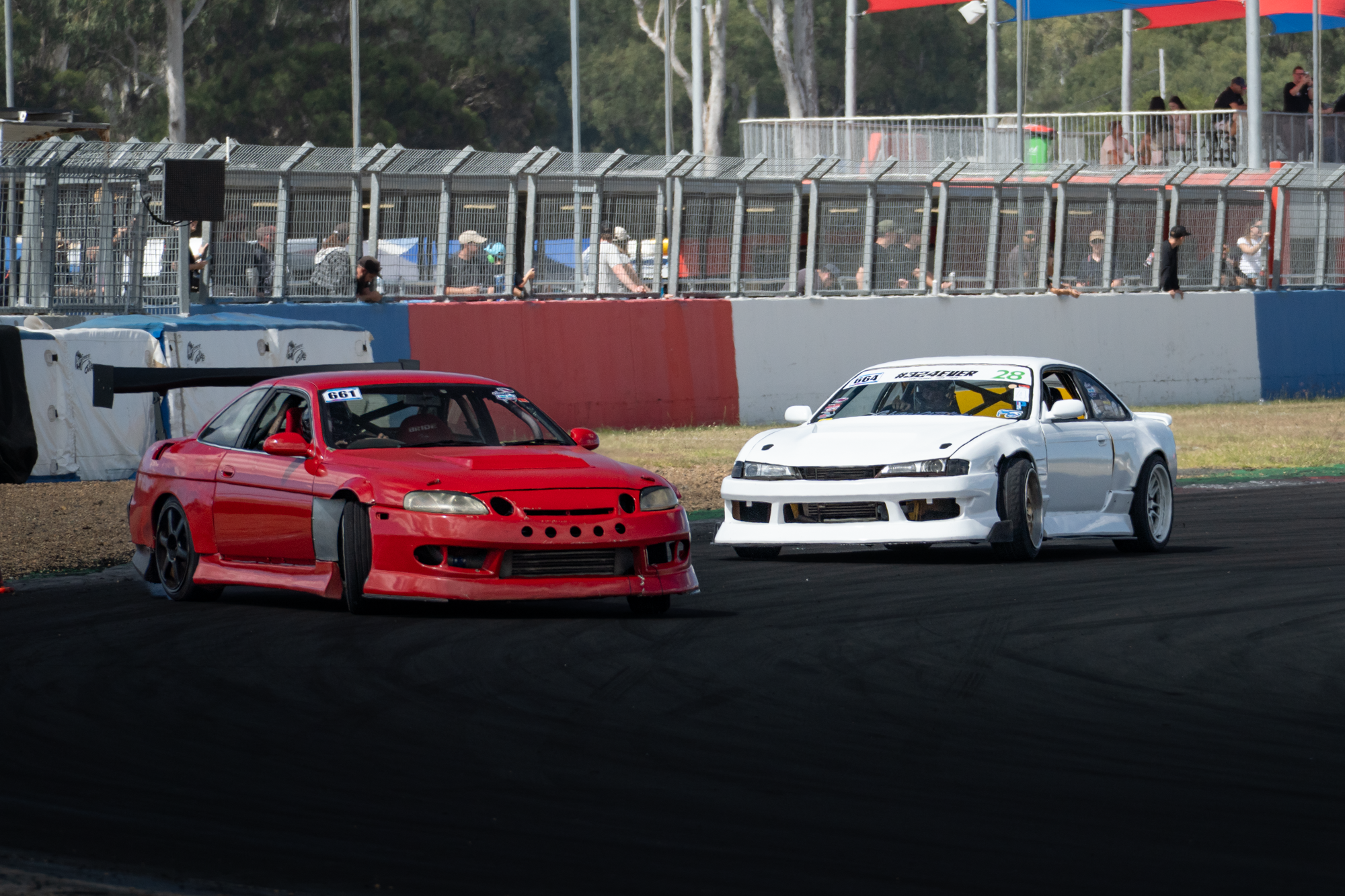 Race cars on a racetrack with spectators behind a safety barrier, trees in the background.