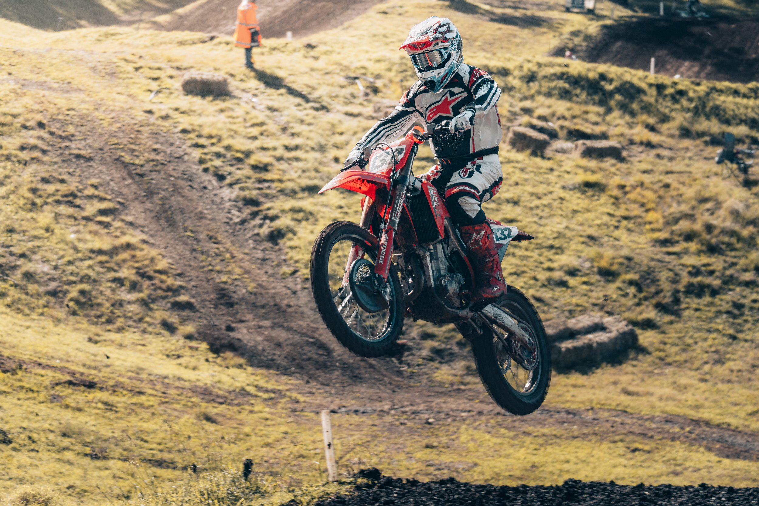 A person wearing a helmet and motocross gear riding a dirt bike through a hilly outdoor terrain with yellow grass, with a person in an orange vest and other motorcyclists visible in the background.