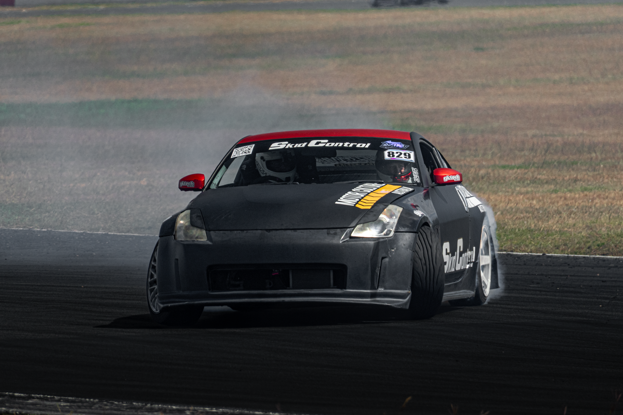 A black race car with various sponsor stickers, including 'Skid Control,' is drifting on a racetrack, kicking up smoke as it turns sharply with a driver wearing a helmet visible through the windshield.
