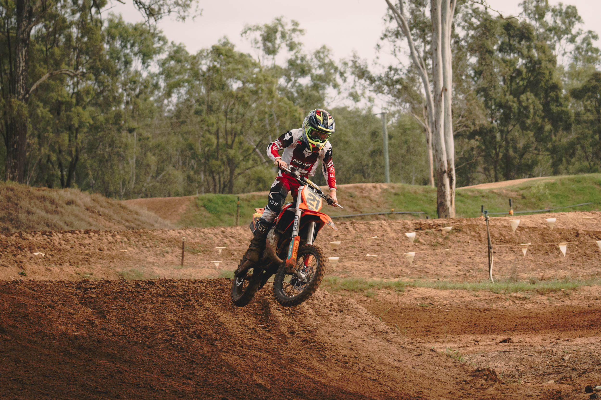 A motocross rider in full gear riding a dirt bike on a dirt track with trees in the background.