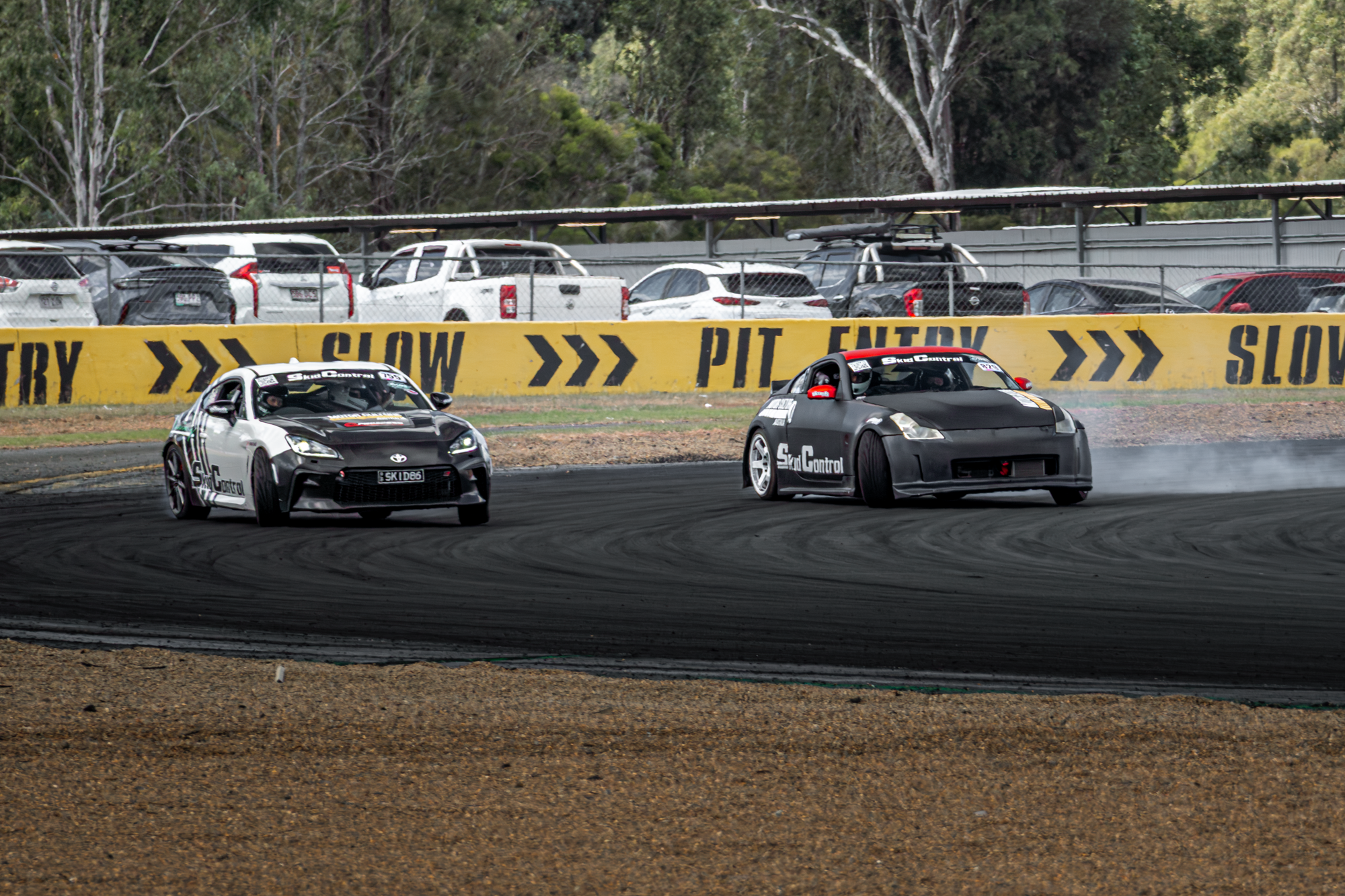 Two race cars on a dirt track, one black and the other grey, with tires kicking up dust; a yellow barrier with black text saying 'SLOW PIT ENTRY' and a chain-link fence in the background, along with parked cars and trees.