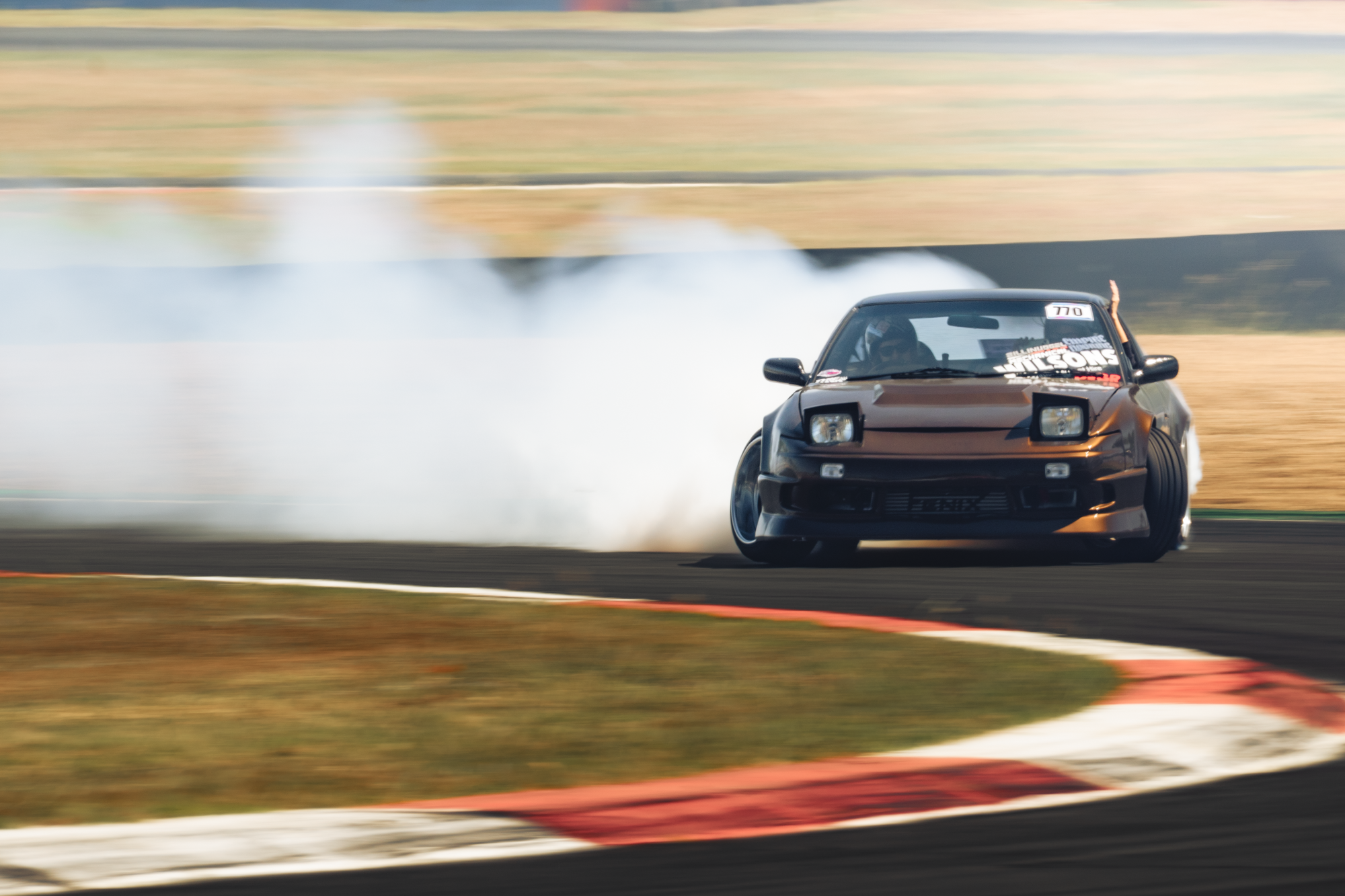 A black sports car drifting on a race track, creating a cloud of tire smoke, with a blurred background of grassy fields and a blue sky.