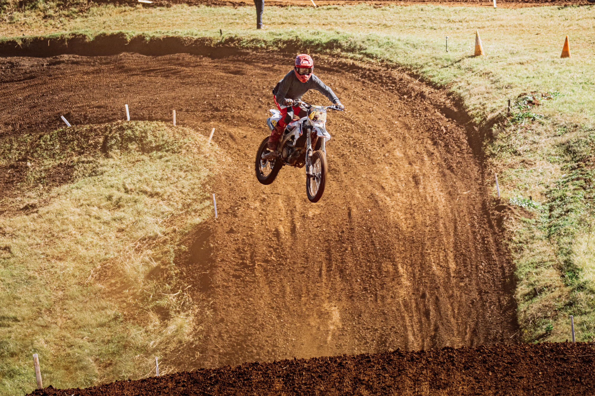 A motocross rider wearing a red helmet, black gear, and red pants riding a dirt bike on a dirt track, surrounded by grassy areas with orange cones, during daytime.