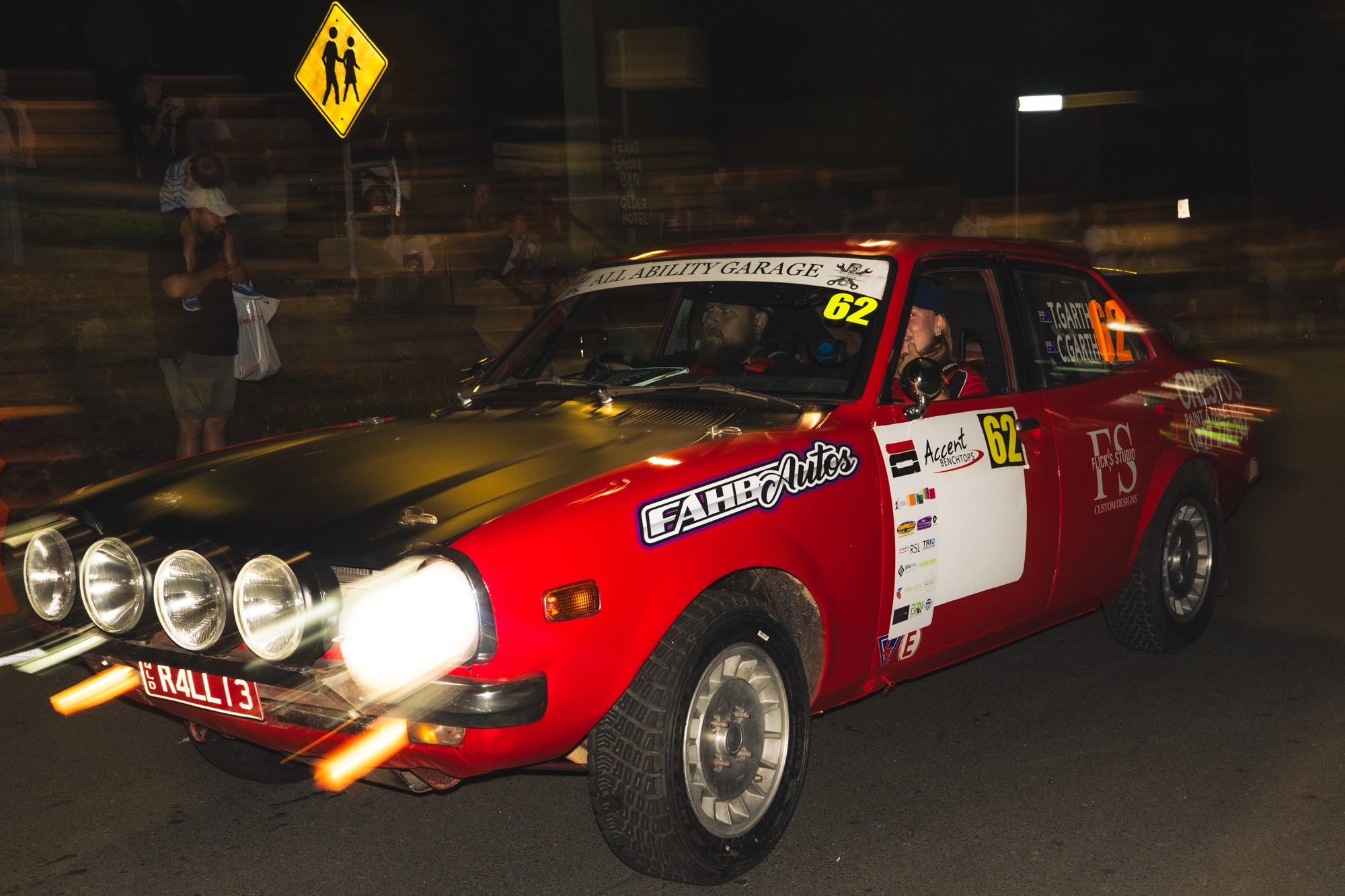 A vintage red rally car with multiple front lights, racing number 62, and various sponsor stickers on the side. It's on a dark street at night with motion blur, and there is a yellow pedestrian crossing sign in the background. Several people are stan
