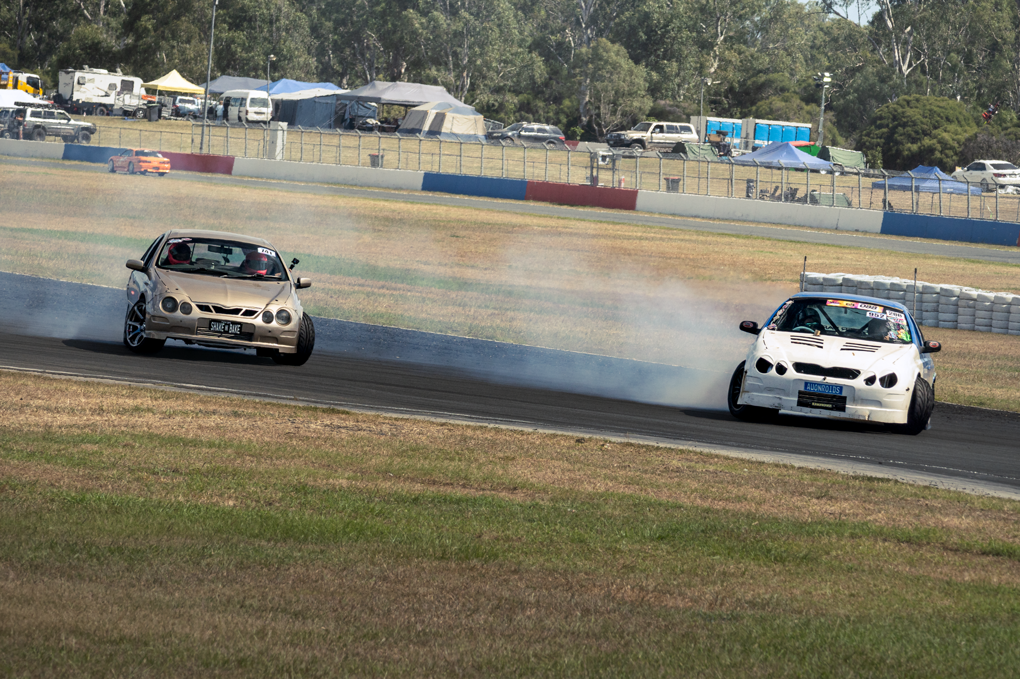 Two cars racing on a dirt track, kicking up dust, with a spectator area and tents in the background.